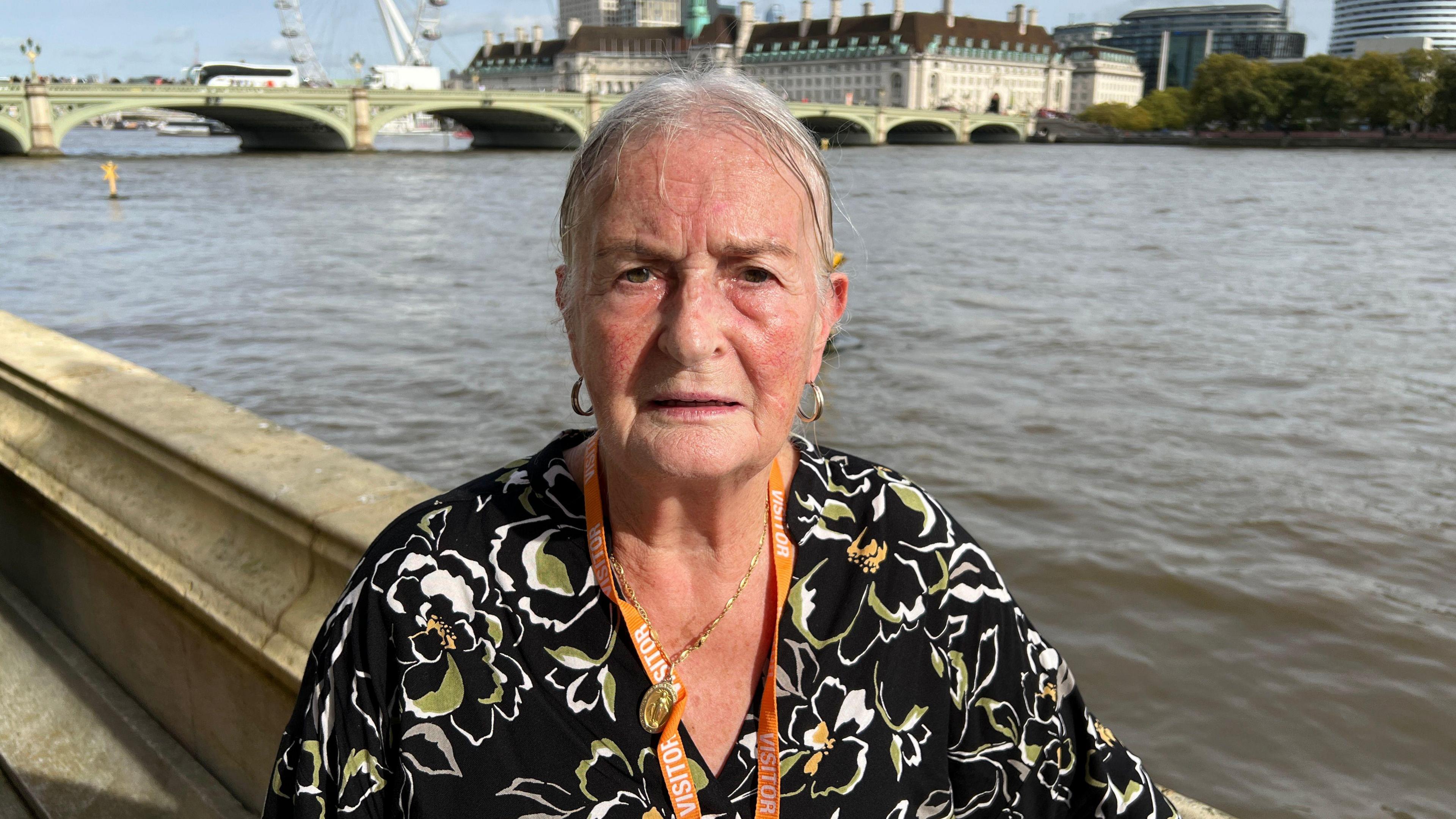 Big Communi-Tea client Jackie Carroll is standing on the House of Commons terrace in front of the River Thames. The London Eye wheel is in the background. She is wearing a black blouse with gold and white flowers and a gold necklace.