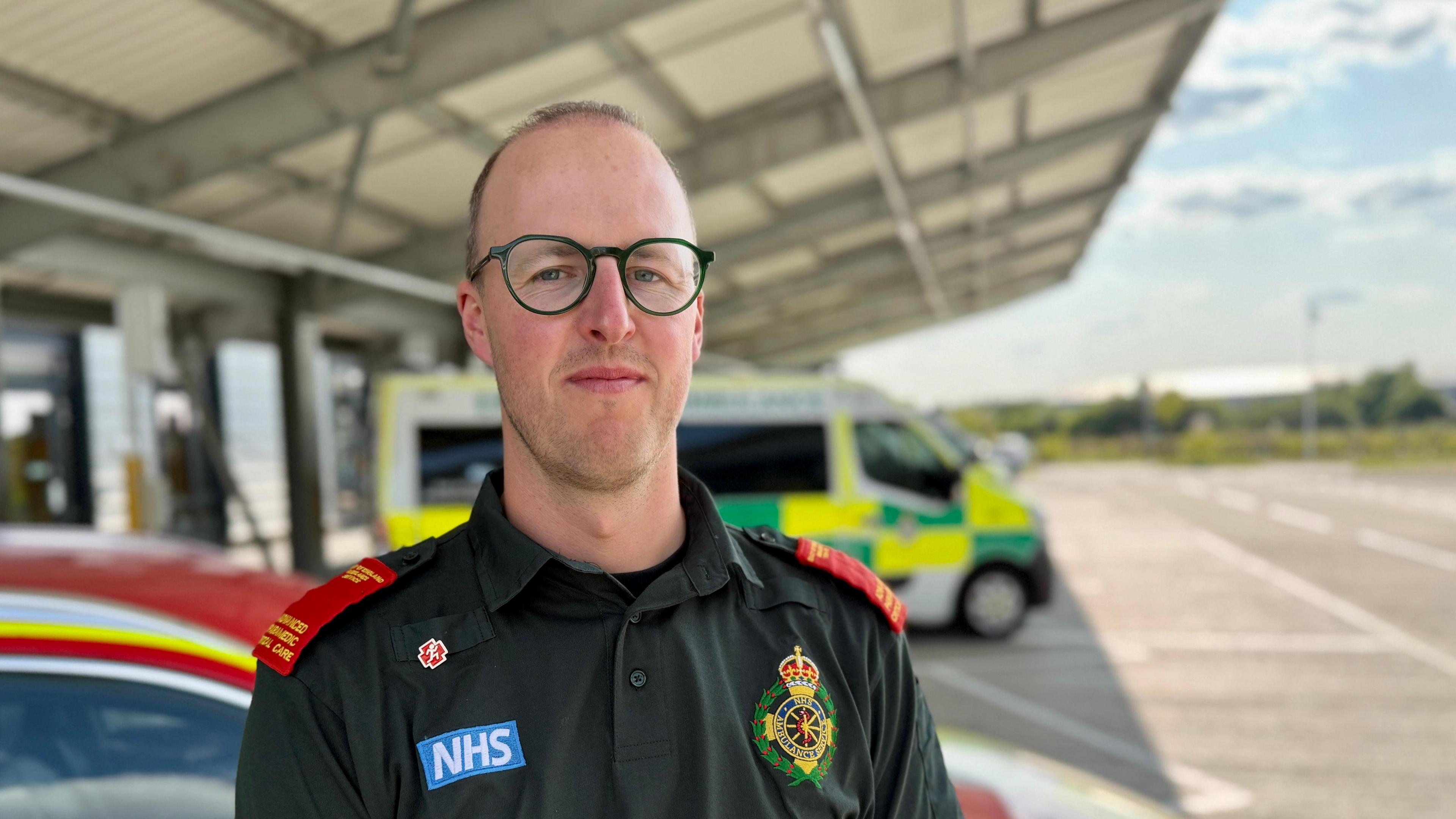 Advanced paramedic Liam Sagi outside the ambulance station at Bury St Edmunds. It is newly built and has a winged roof. Mr Sagi is wearing a green uniform with an NHS badge and ambulance service crest. He has red stripes on his shoulders. There is an ambulance parked in the background.
