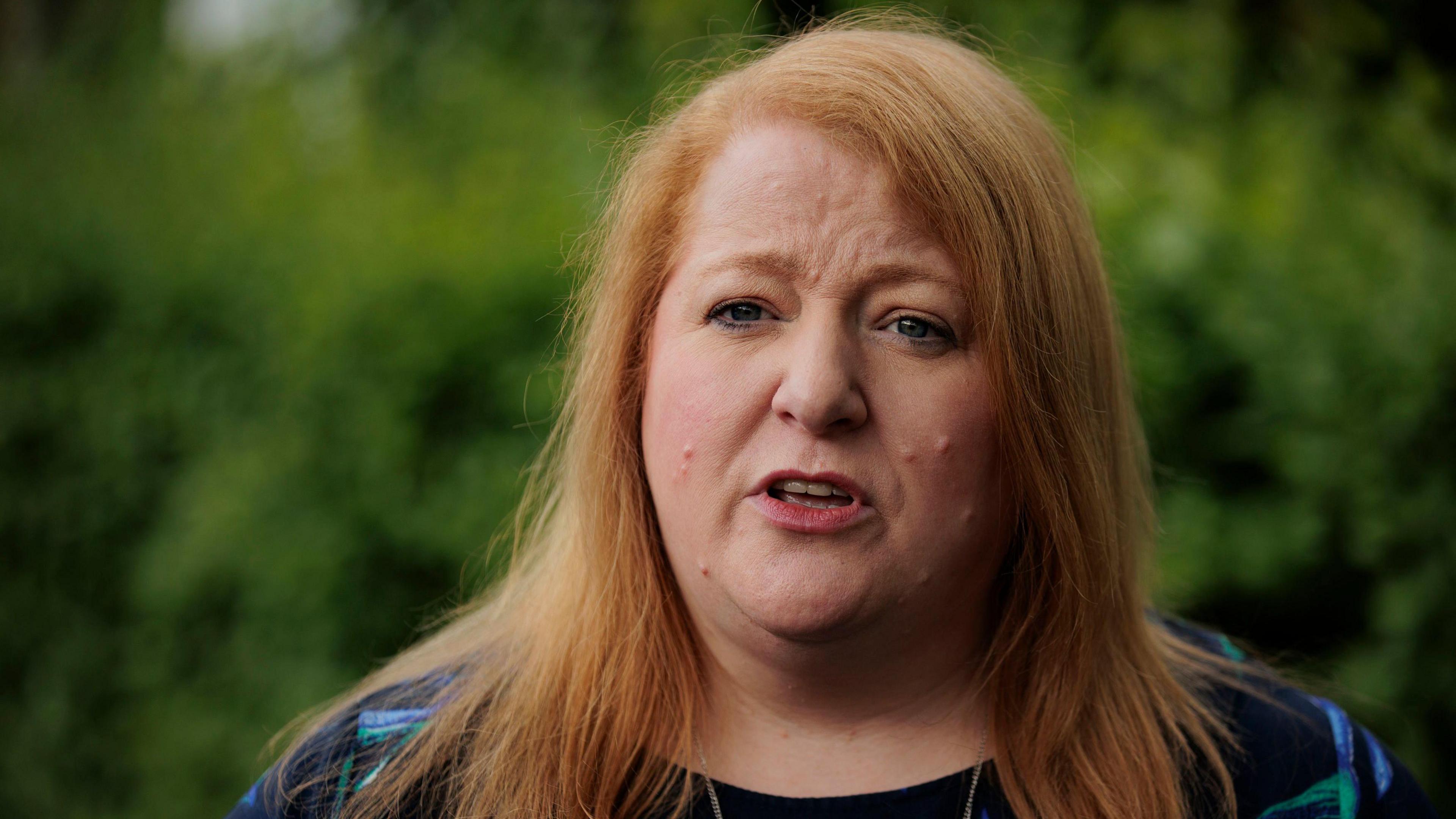 Headshot of Naomi Long looking at the camera a speaking with her eyebrows furrowed. Behind her some greenery that is out of focus.