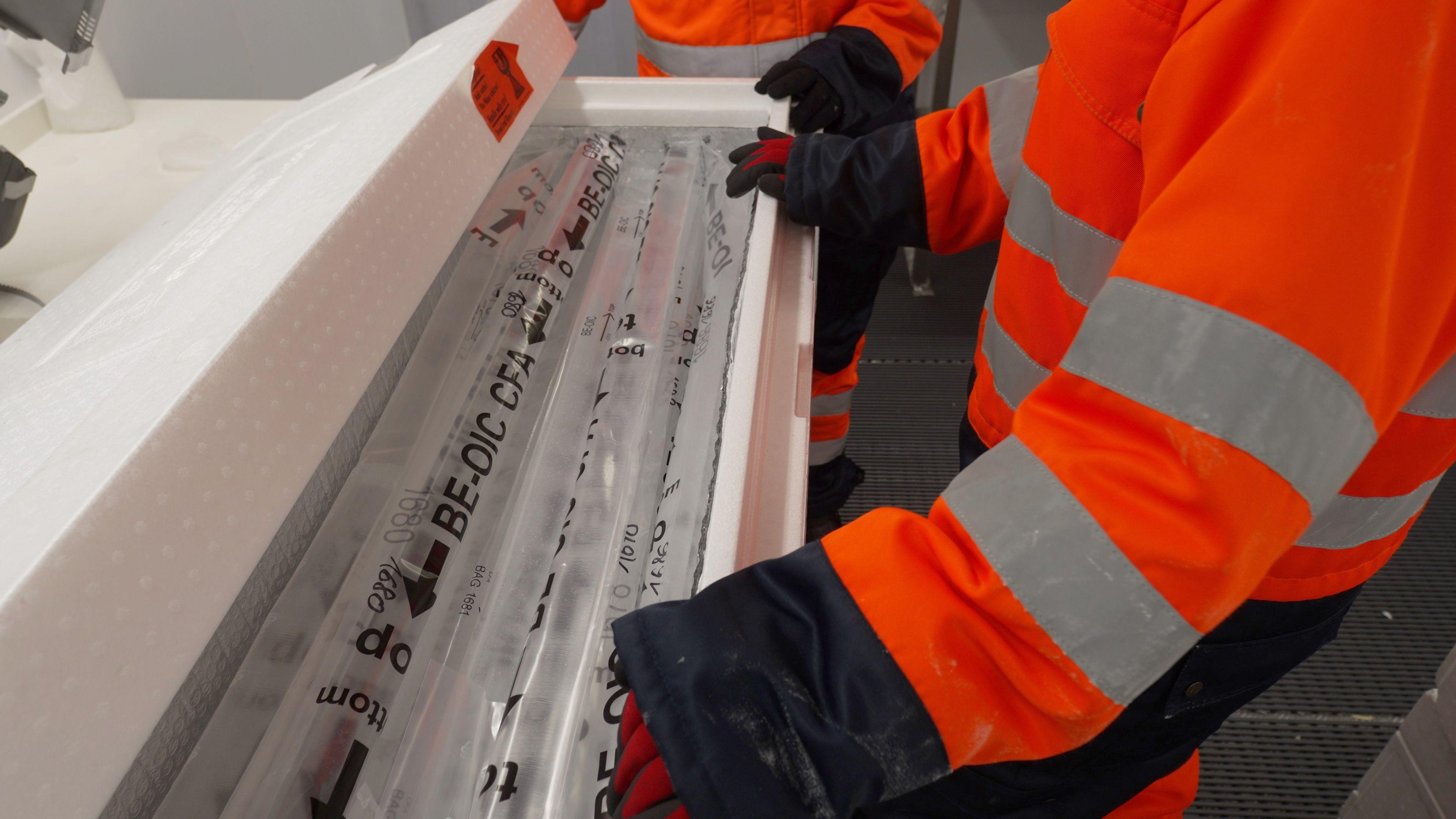 A person wearing orange and black protective clothes and gloves has their hands over long tubes of ice covered in plastic wrapping. The tubes are inside a large box.