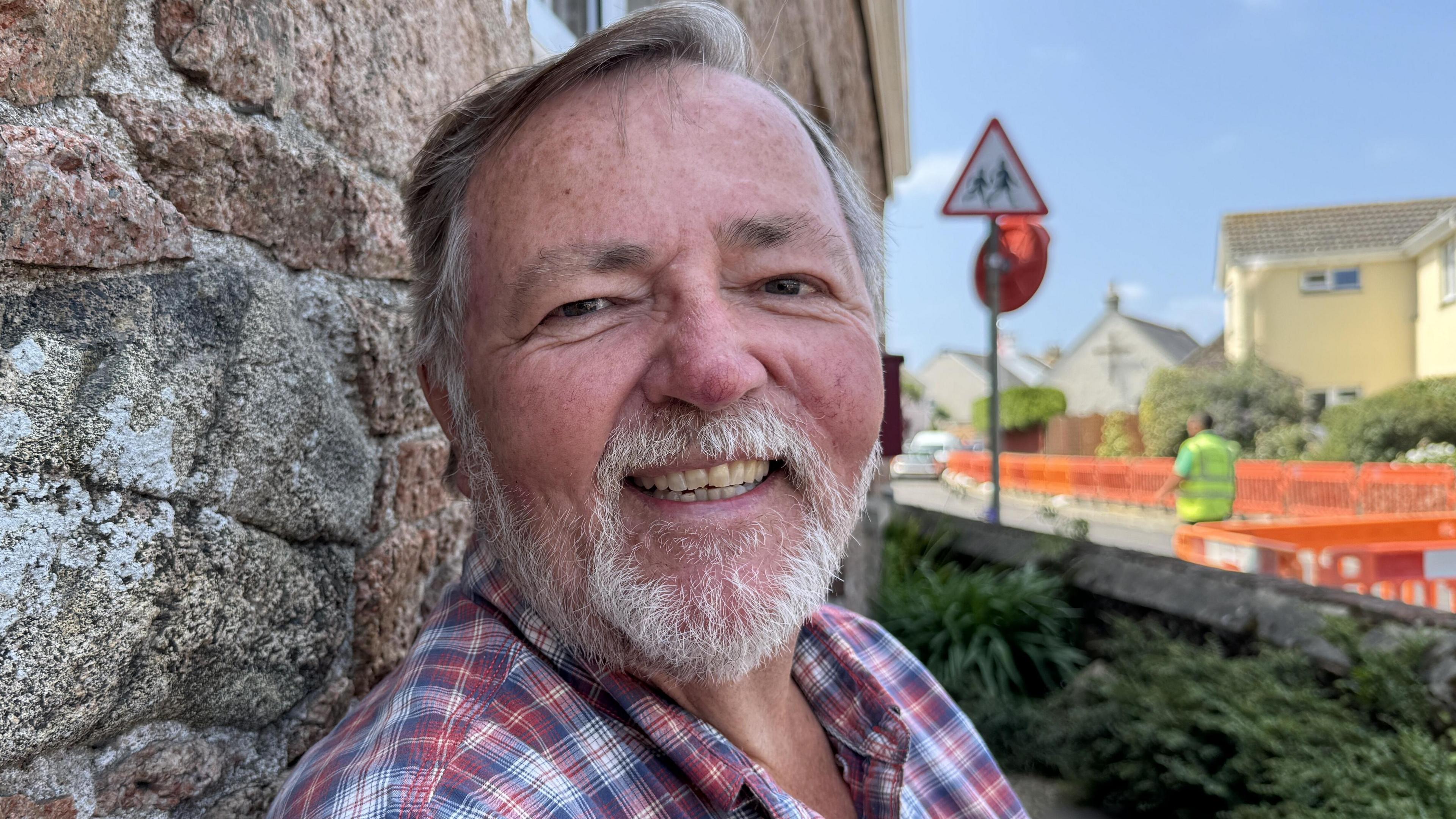 A man is seated against a stone wall. He is wearing a red and blue plaid shirt. Behind him, a street scene includes orange construction barriers and a triangular caution road sign. Further in the background, residential houses are visible beneath a mostly clear blue sky with a few scattered clouds.