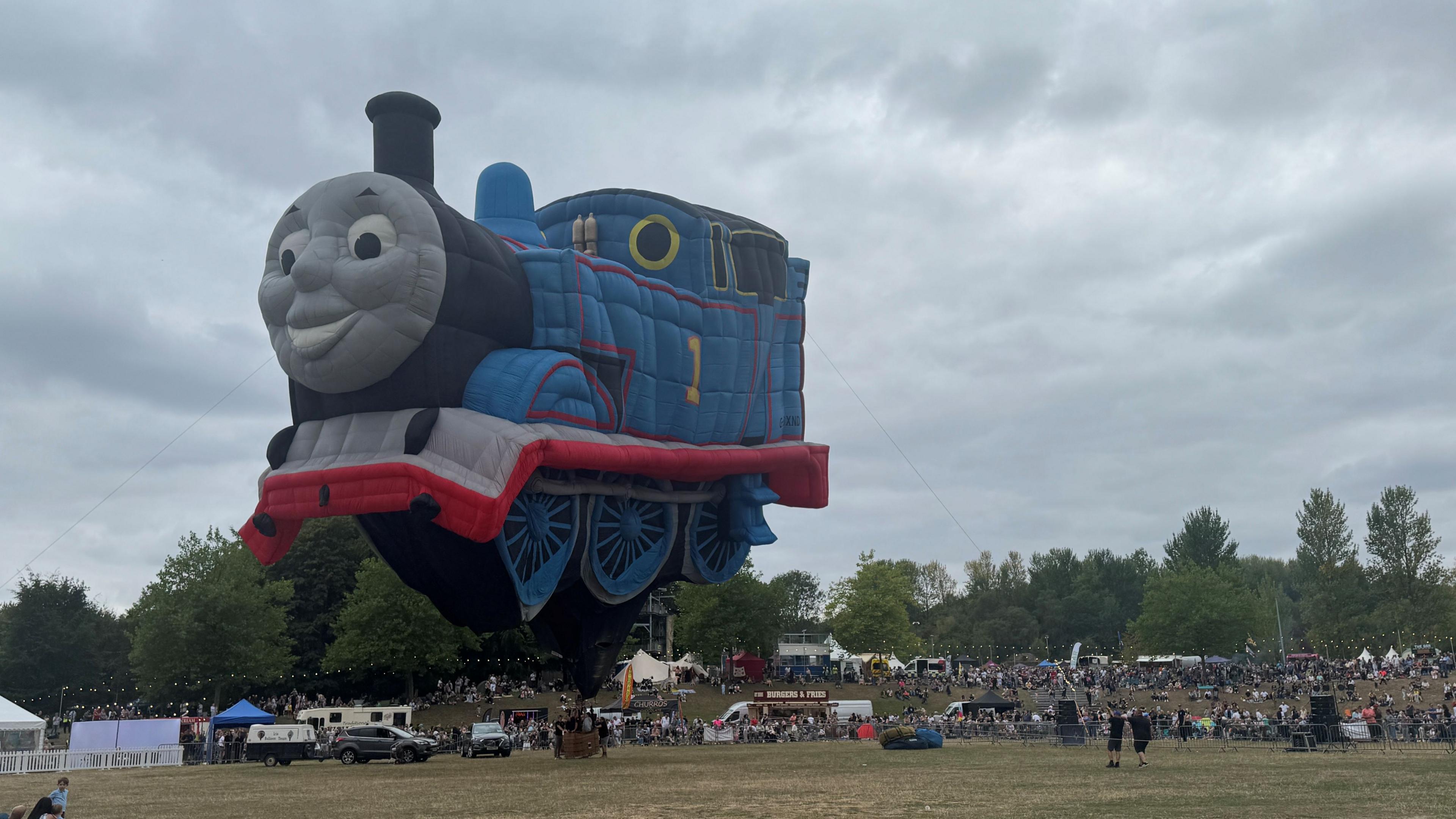 A large Thomas the Tank engine balloon tied down with rope. It is blue, grey and red with a grey face. Stalls and vehicles and some people are also in the picture.