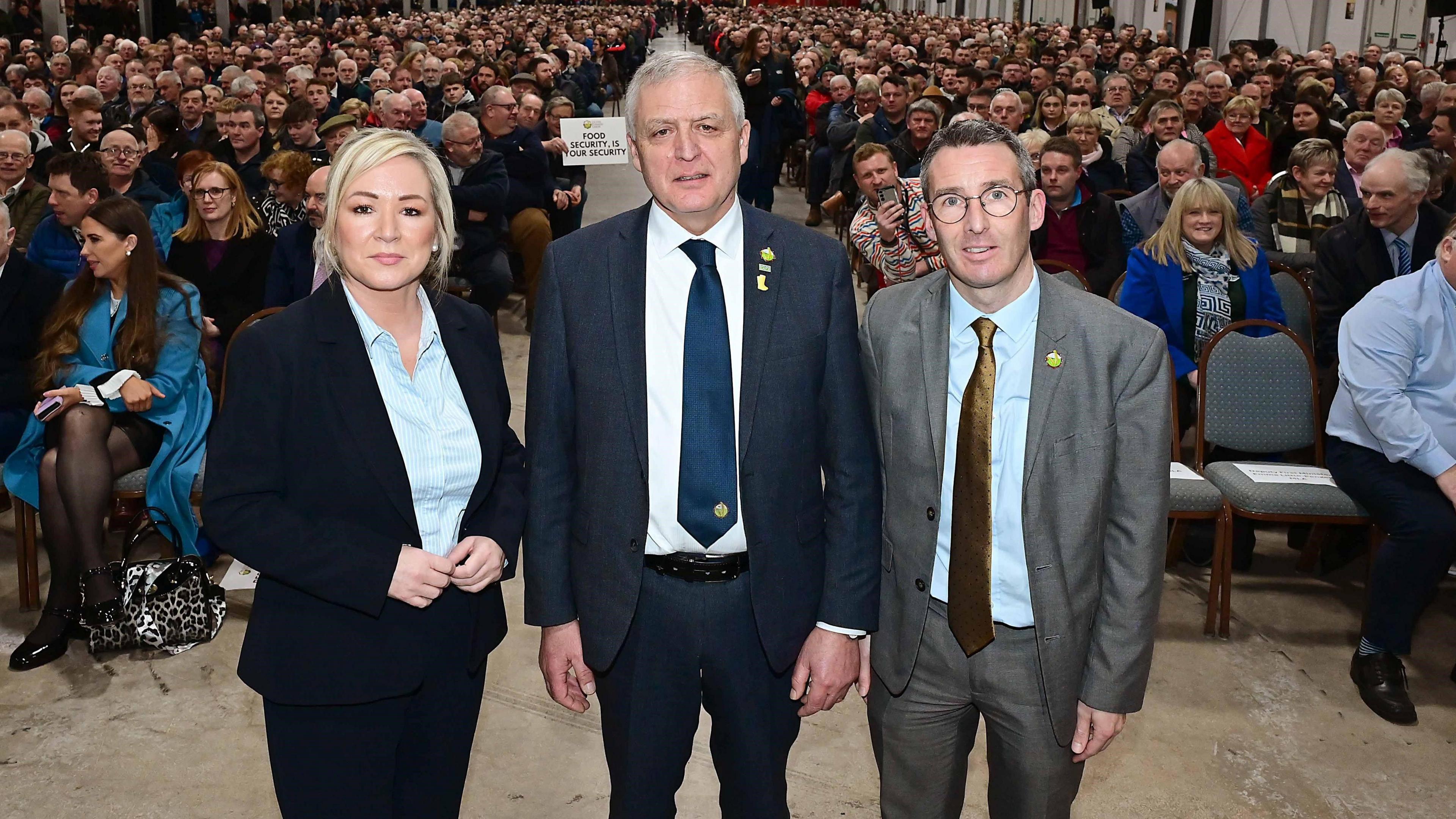 First Minister Michelle O'Neill, dressed in a navy trouser suit and blue shirt, on the left, with Agriculture Minister Andrew Muir, dressed in grey suit and brown tie, on the right. In the middle is William Irvine in a navy suit and navy tie. He has grey hair. The exhibition is full of people sitting on chairs.