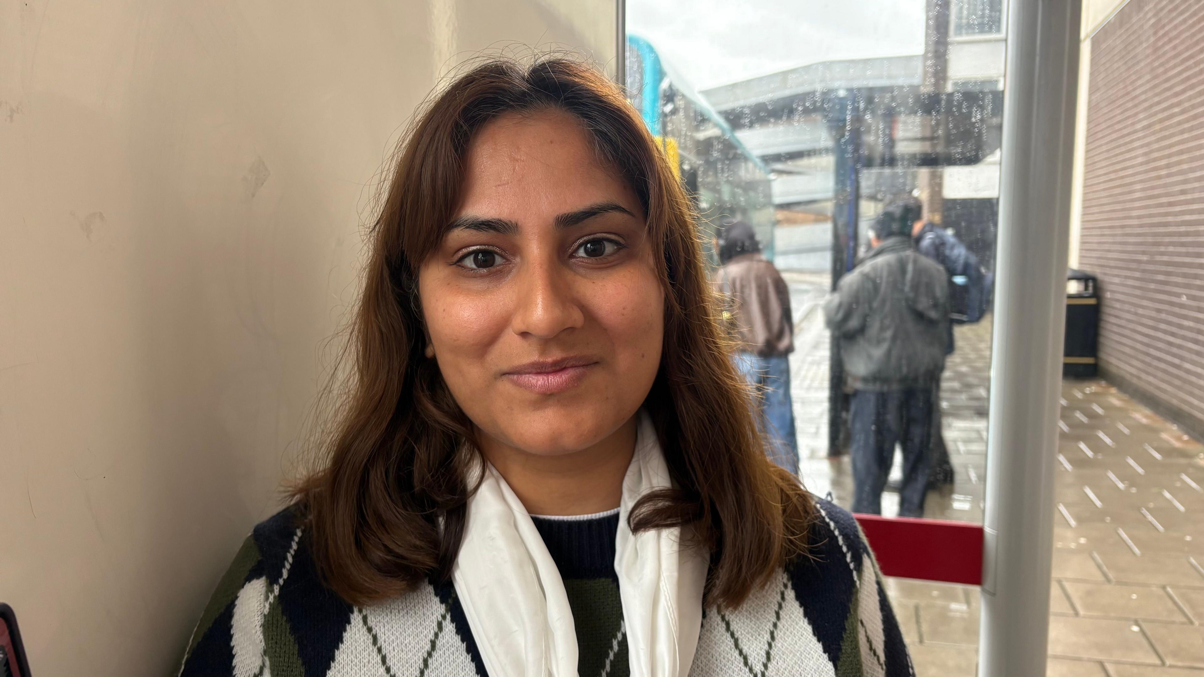 Iqra Bashir with long dark brown hair smiling at the camera and wearing a black and white top. She is in a white-walled bus shelter and people are visible getting on to a bus behind her.