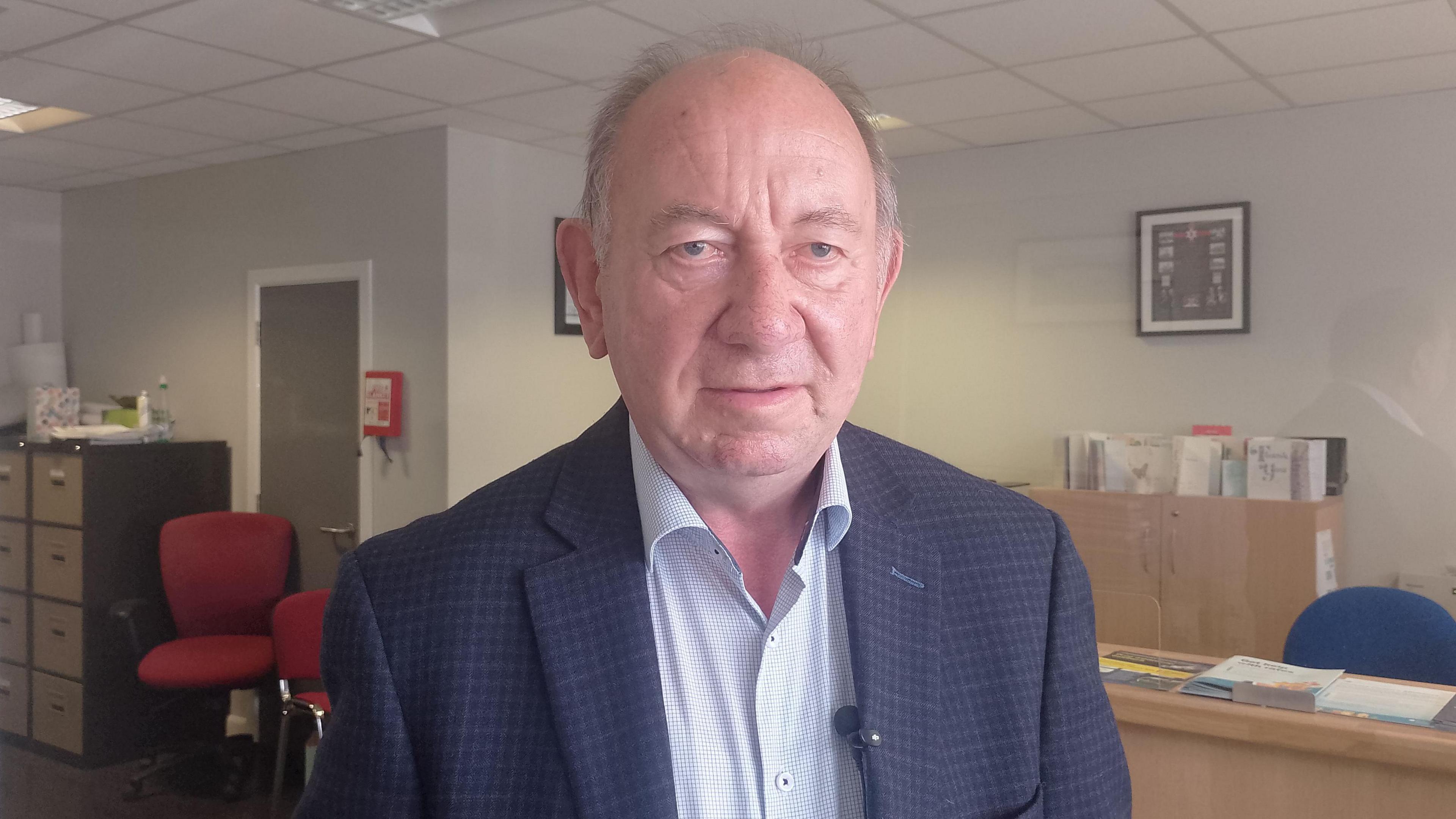 William Irwin is wearing a navy, checked suit jacket over a pale coloured shirt. He has grey hair and is standing in an office.