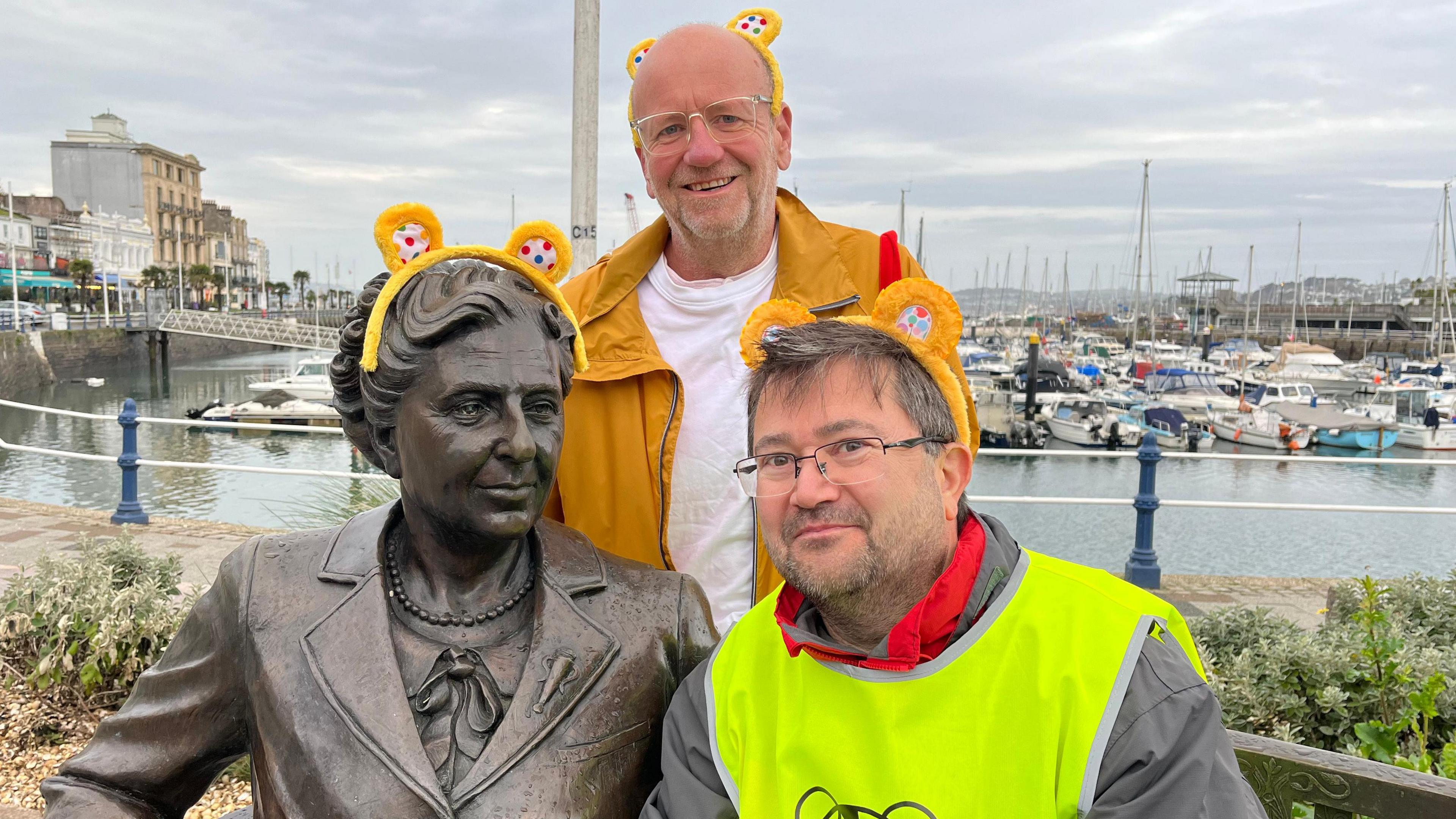 BBC Radio Devon presenter Michael Chequer and Matt Newbury from the Agatha Christie Festival are next to the Agatha Christie bench statue on Torquay harbour. The men and statue have Pudsey Bear ear Alice bands on.