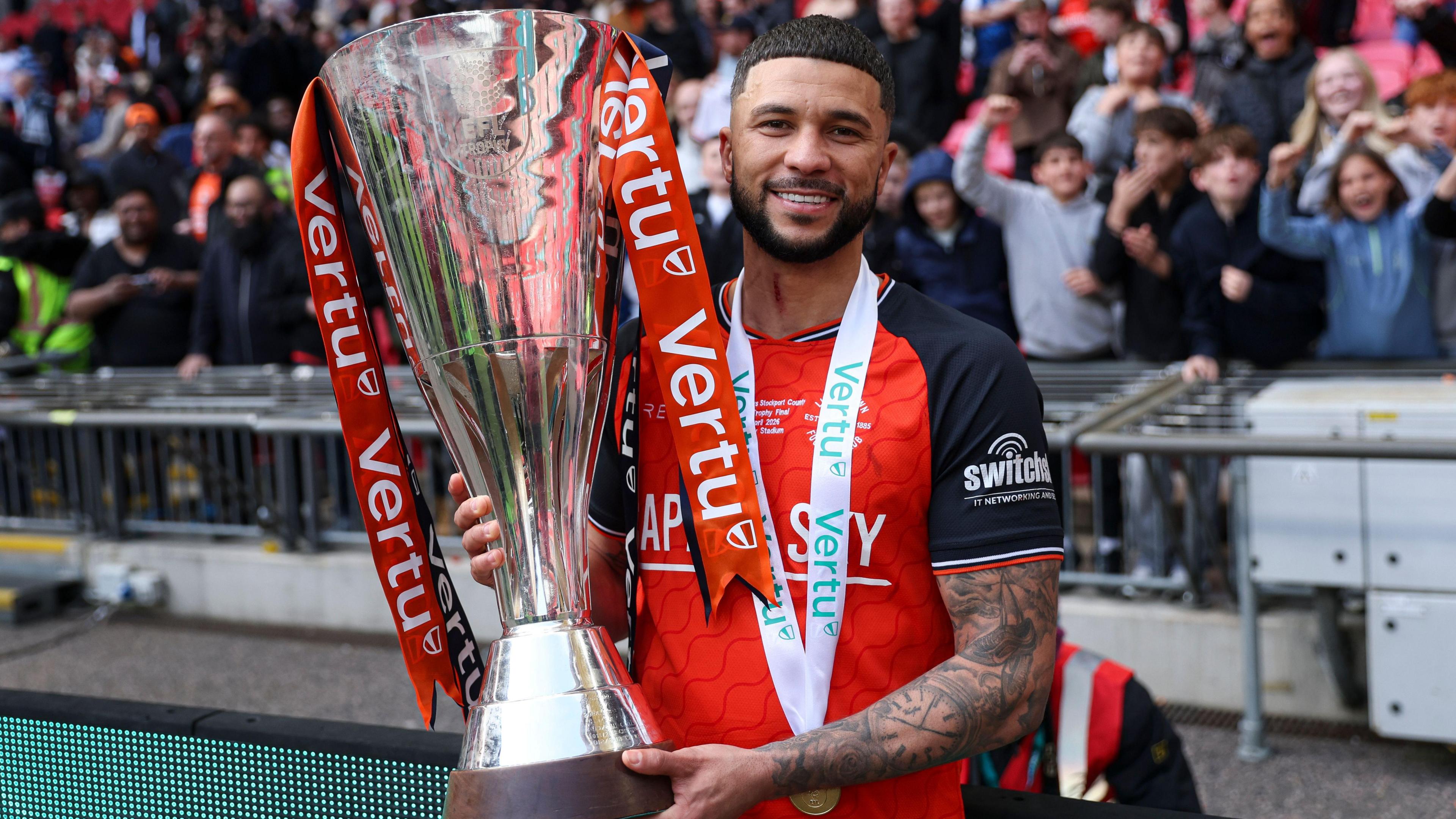 Nahki Wells, wearing Luton's orange home shirt, smiles while holding the Vertu Trophy