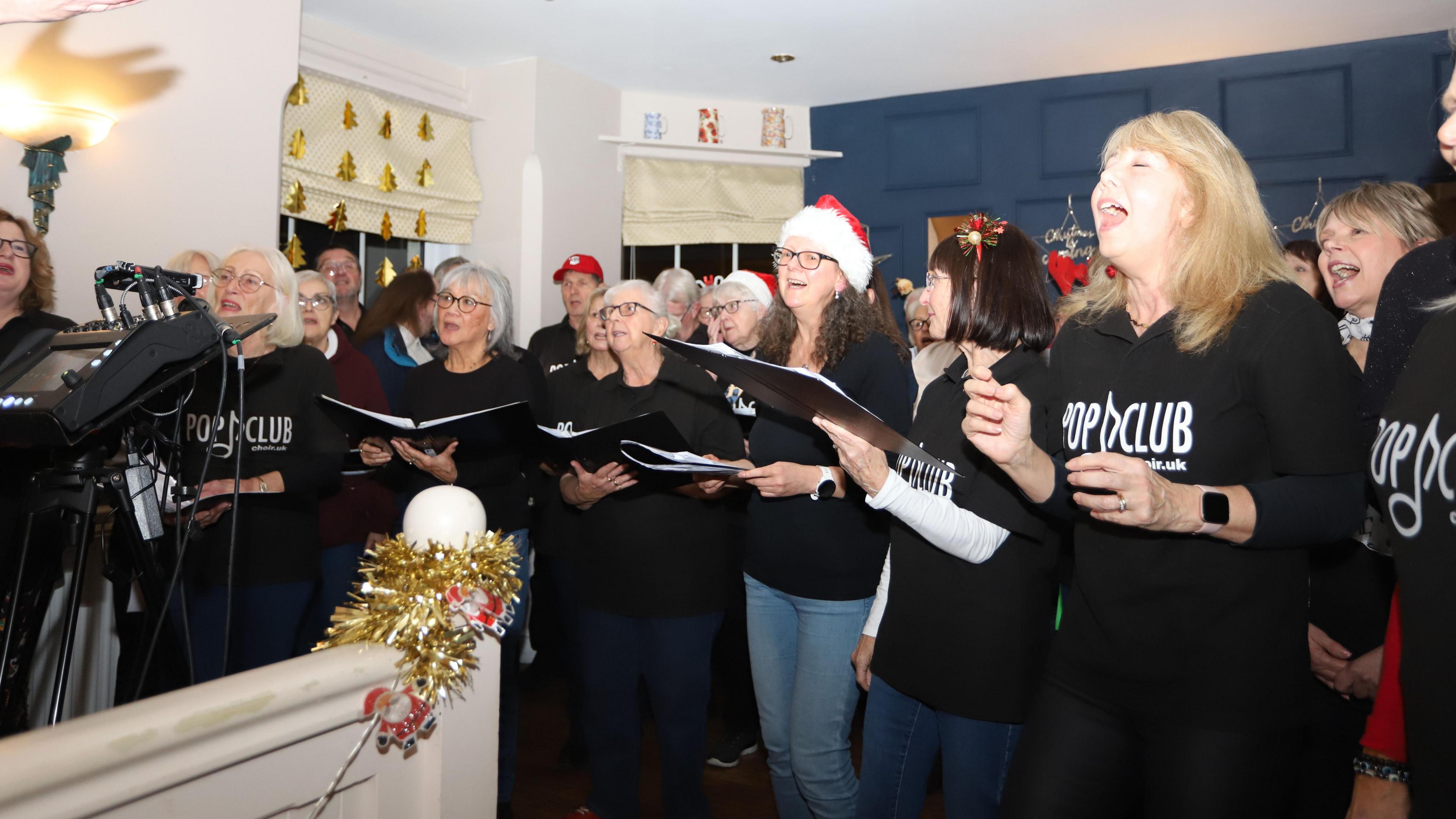 A choir is singing from song sheets in a room with a blue wall. There is tinsel wrapped around a wooden post with santa decorations.