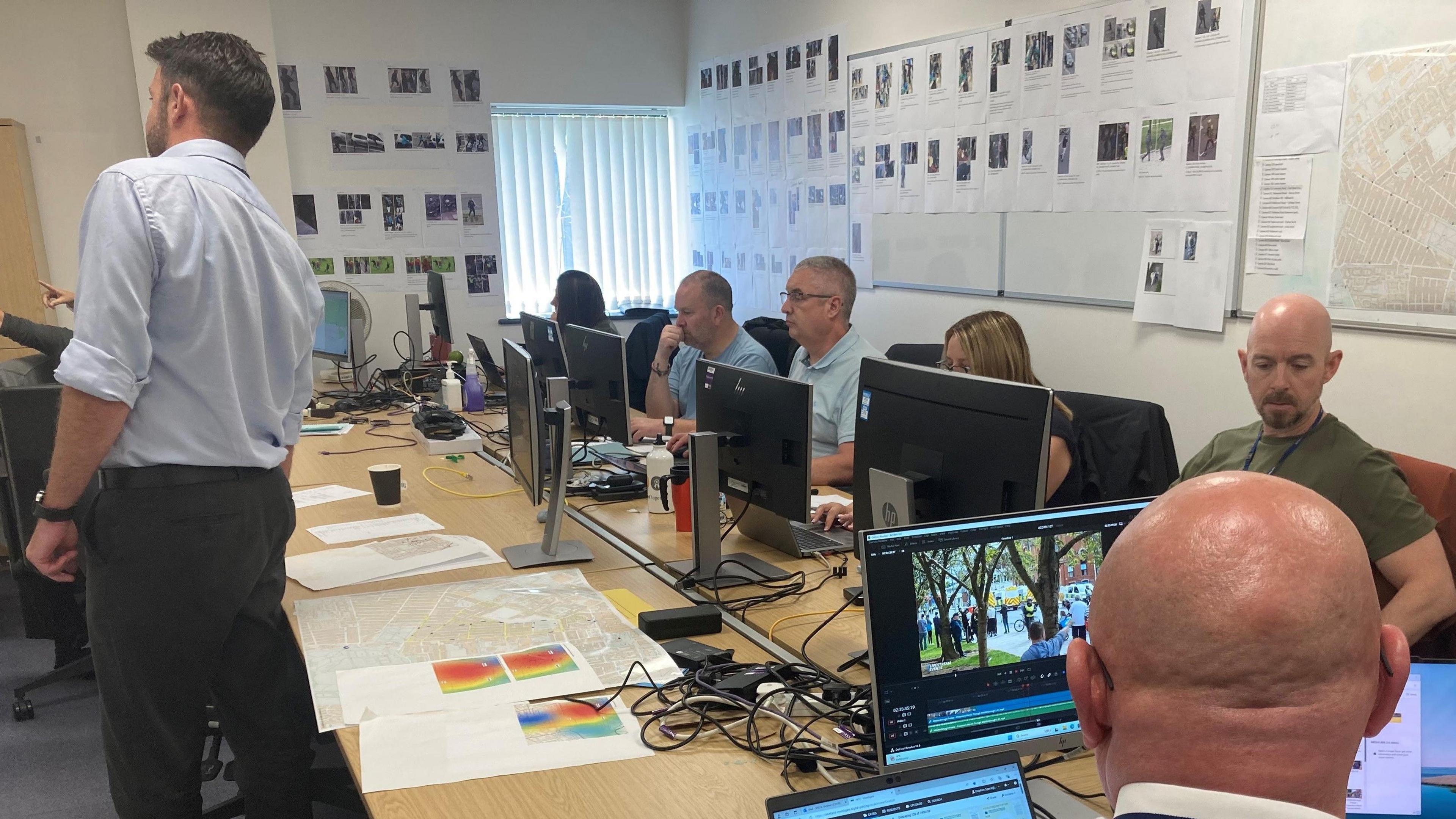 Police incident room, with a standing man looking at one of the walls, which are plastered with documents and photographs. Six other people are seated around a table looking at individual computer screens. As well as the computers there are also large-scale maps on a long table.