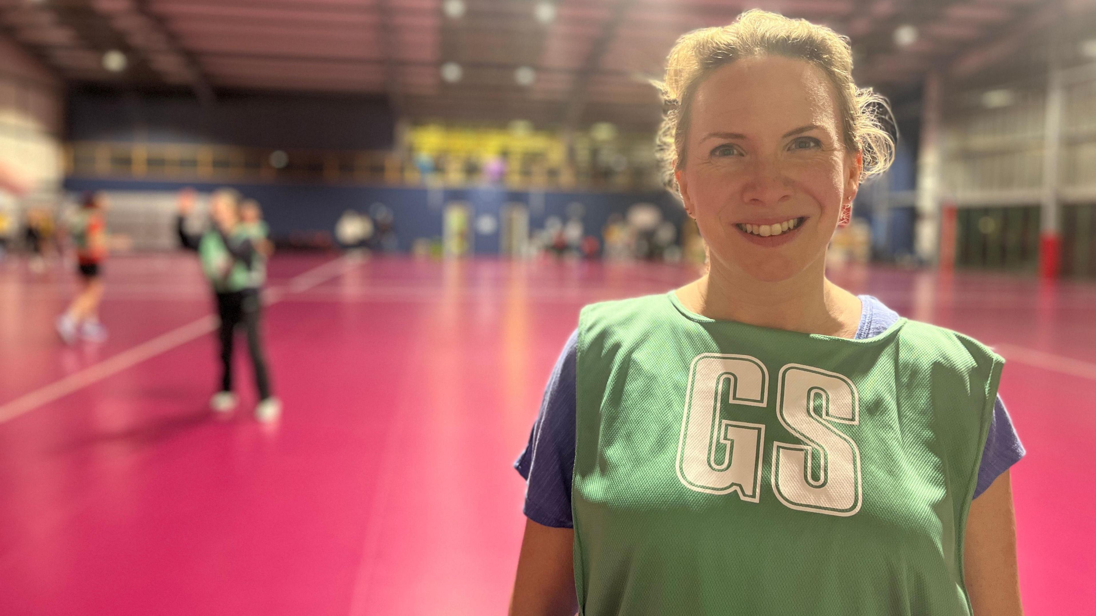 Sarah smiles at the camera as players get ready behind her. She's wearing a blue t-shirt with a light green bib on top with the letters GS on it. She has blonde hair which is tied into a ponytail.