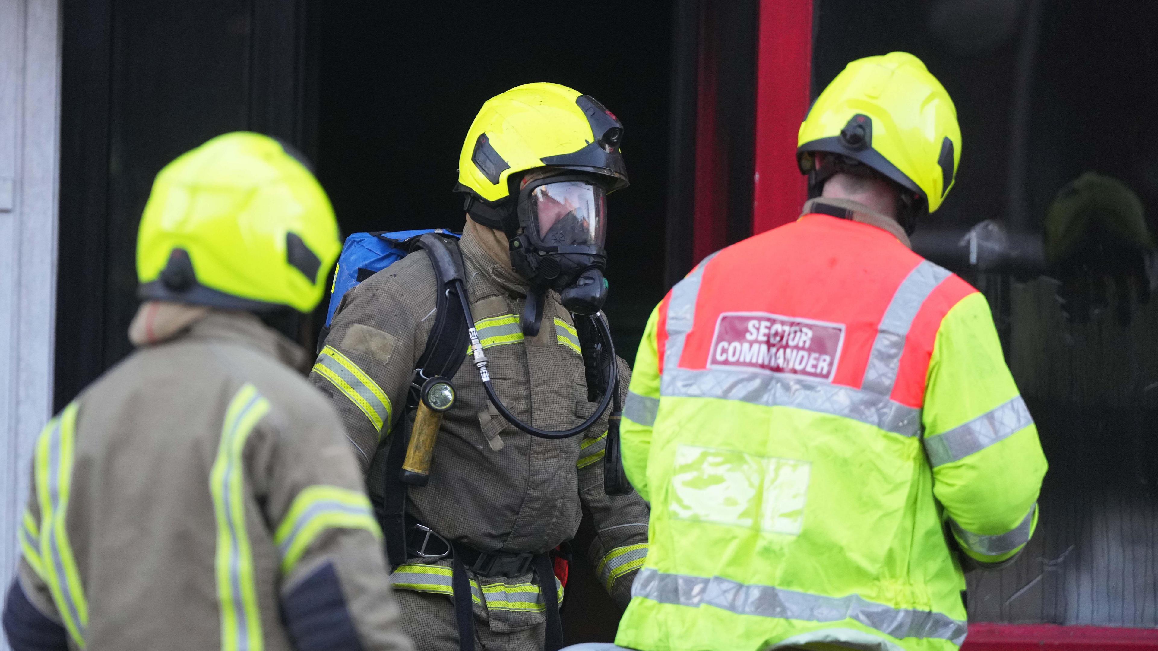 Three firefighters stand together, with one visibly wearing a breathing apparatus. 