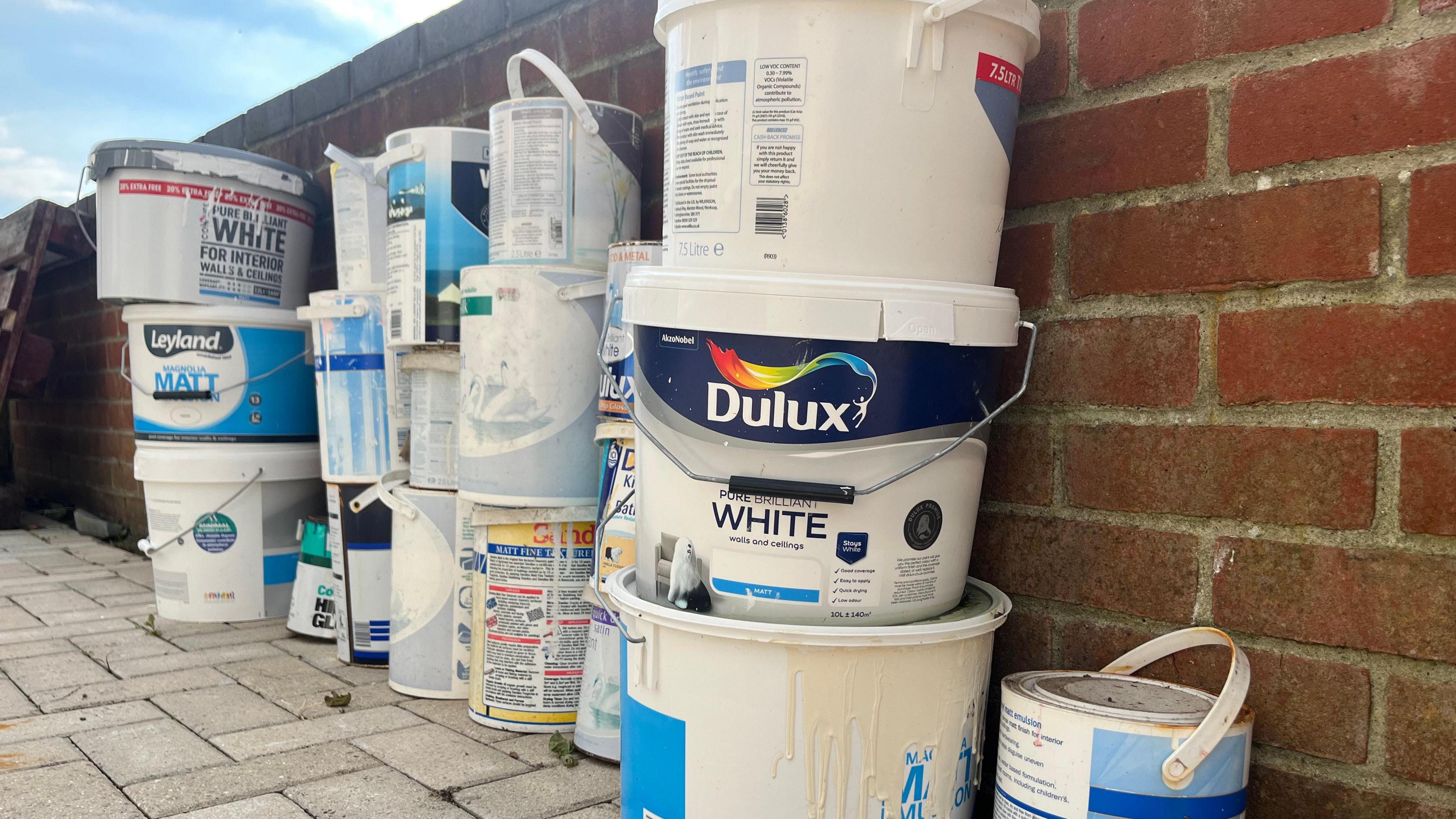 About 19 tins of different branded paint stood next to a brick wall.