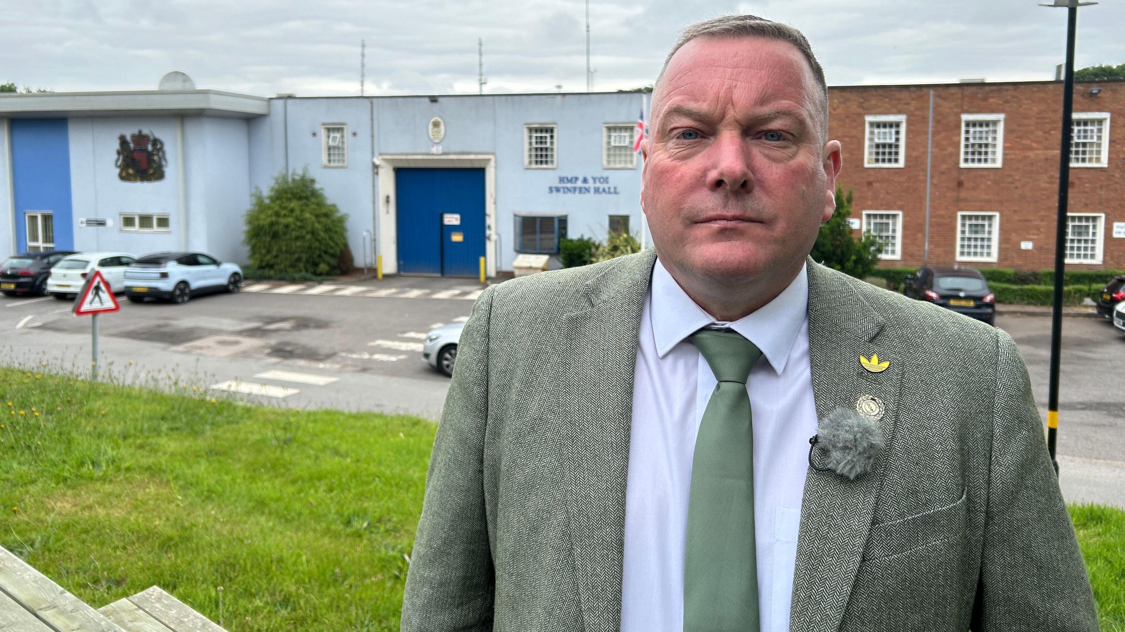 Geoff Willetts of the Prison Officers Association. He is wearing a green suit with some badges on the lapel. He is standing on a hill with Swinfen Hall prison behind him.