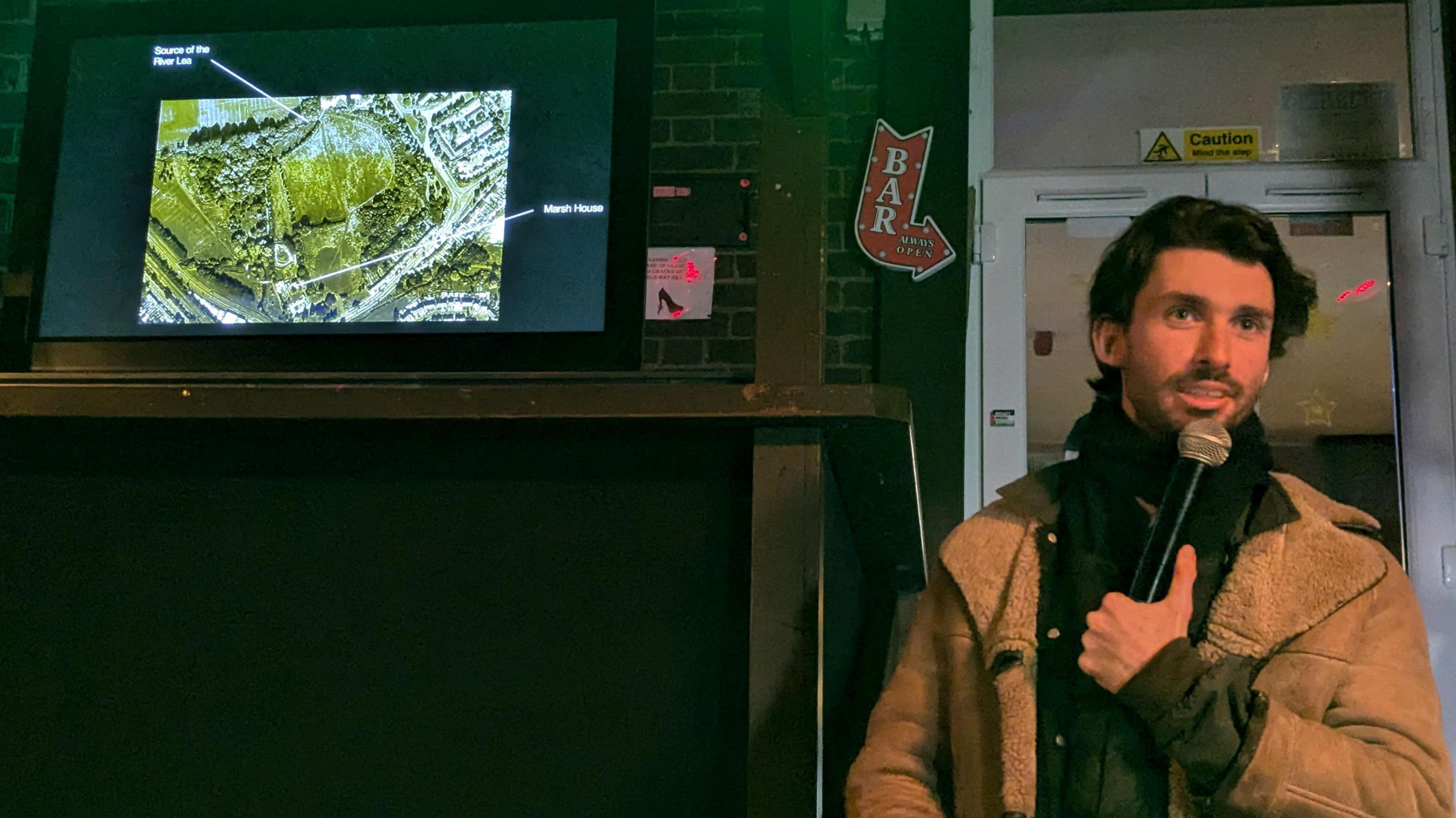 Matthew Rosier, holding a microphone, standing by a television screen that shows an ancient site. He has short dark hair, is wearing a dark top and jumper and camel coloured coat. He is standing to the right of the screen, in front of a door. He is talking. 