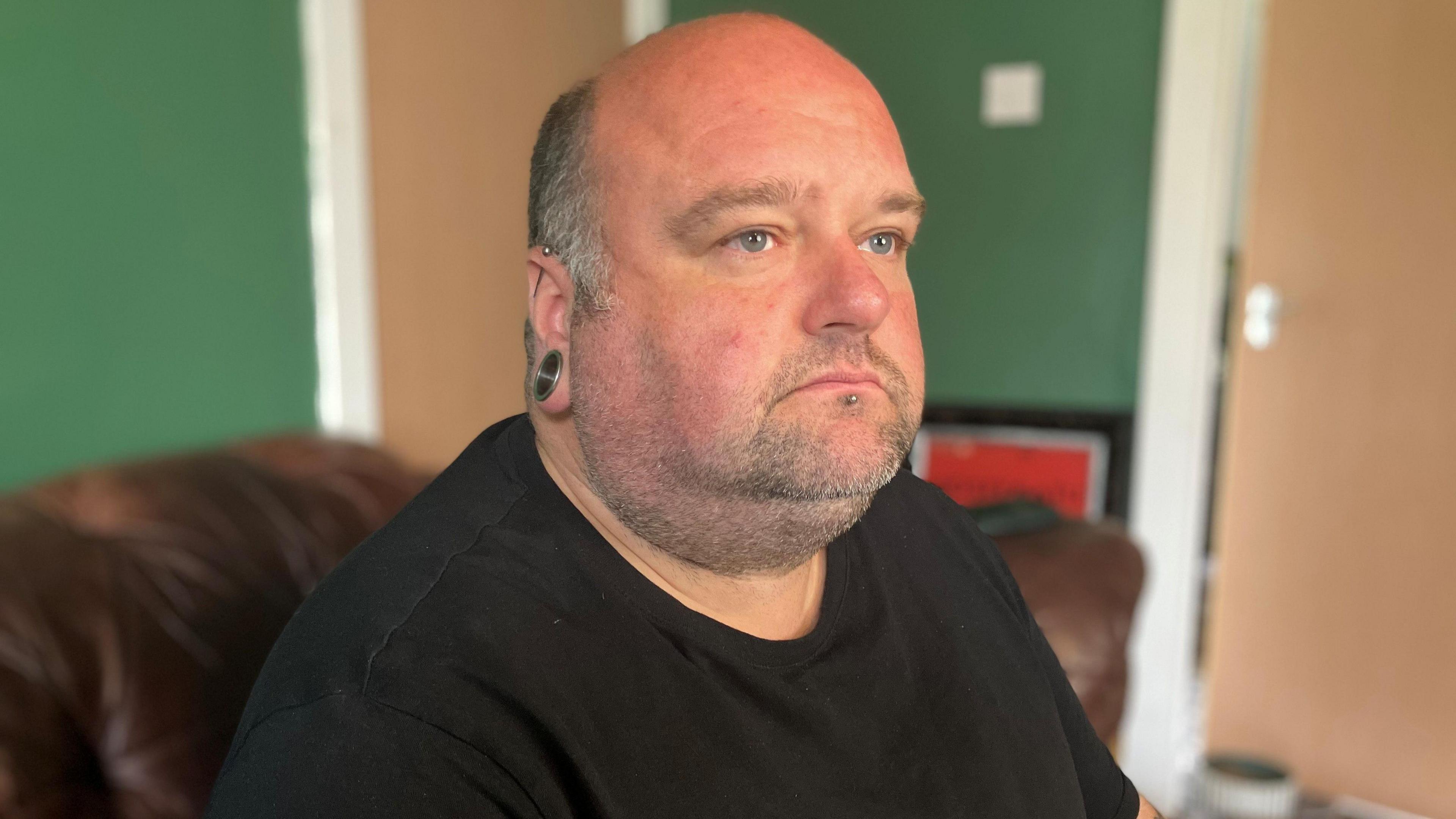 Kevin Doyle sits indoors on a leather sofa looking off tinto the distance. He is a bald man with a large ring in his ear. He wears a black t-shirt