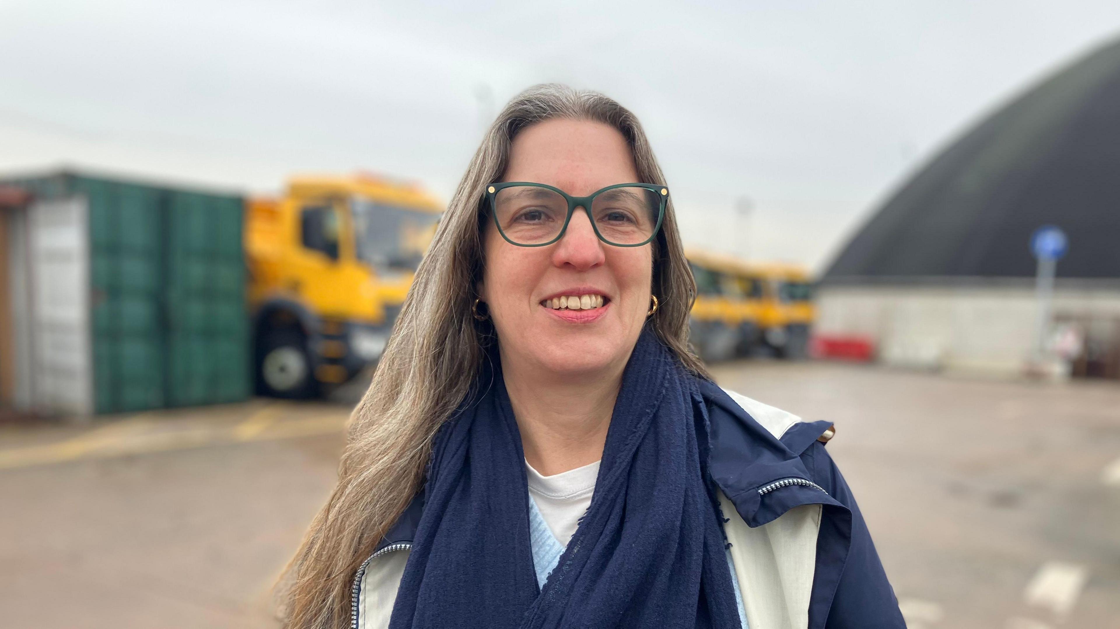 Councillor Laura Robertson-Collins - she has green glasses and a blue and cream coat and long blonde hair. she is wearing gold earrings. you can see a line of gritters behind her. 