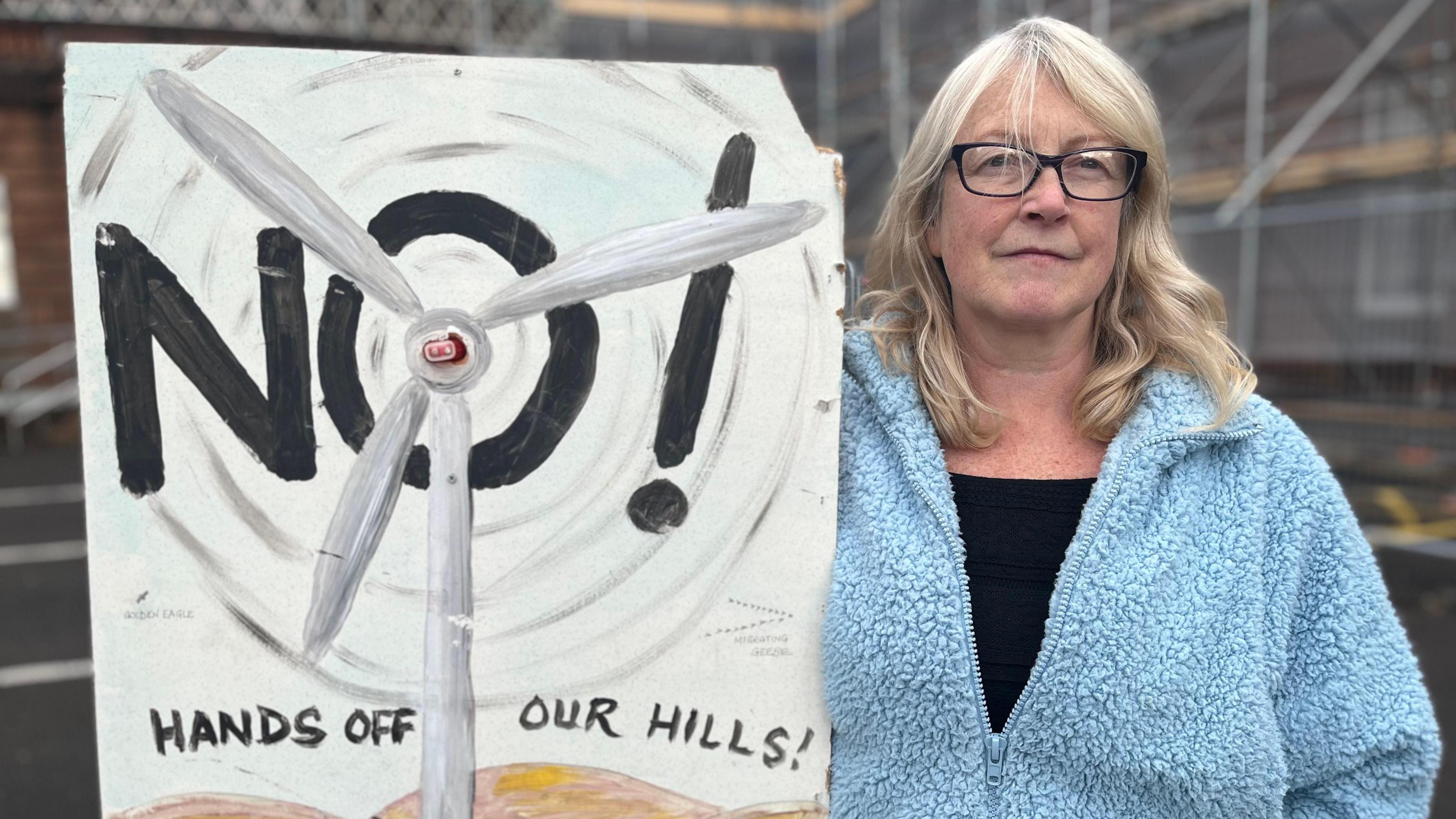 A woman in a light blue fleece top and black T-shirt with long blonde hair and glasses holds up a "Hands Off Our Hills" placard