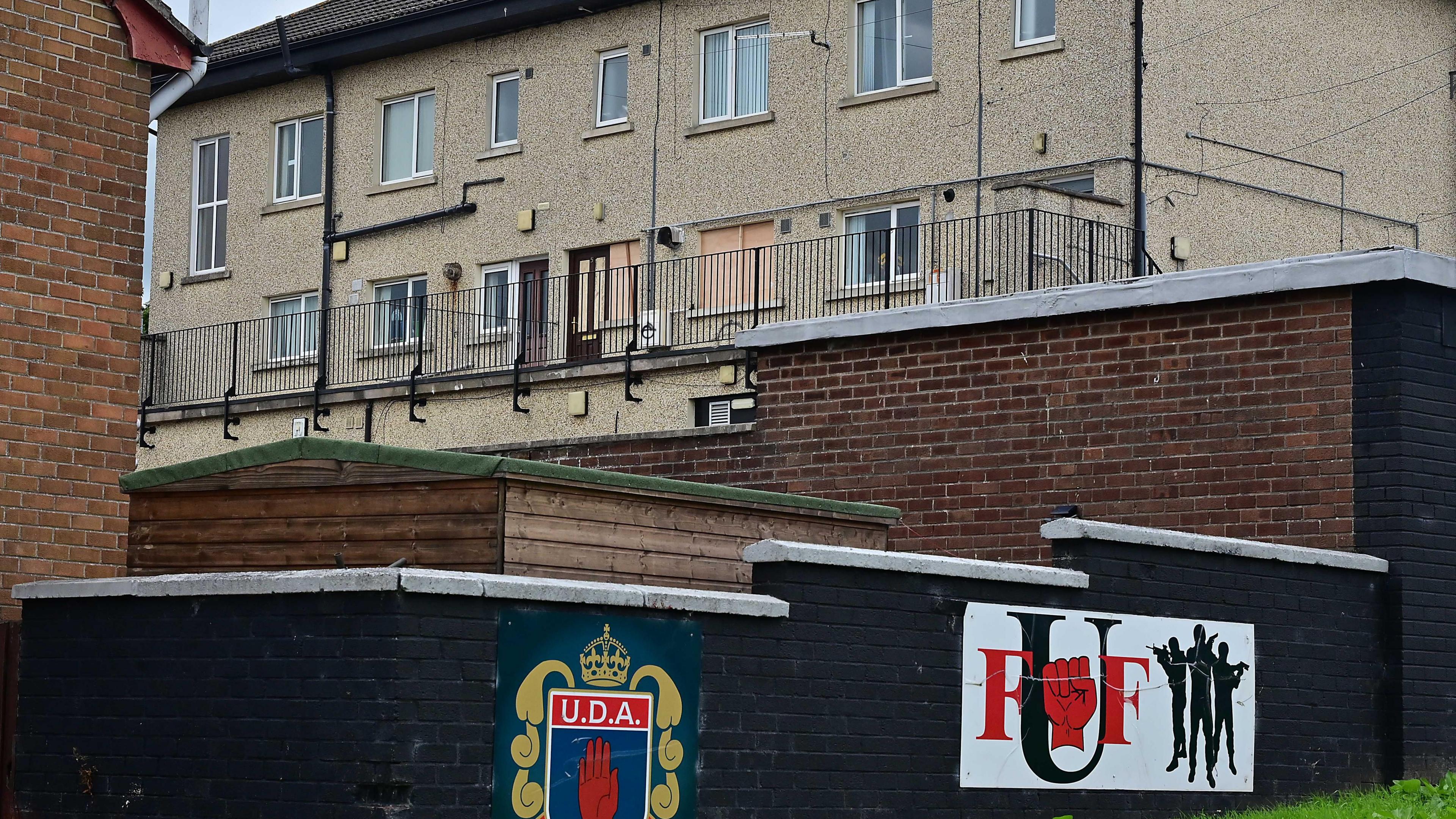 An apartment block rises above a brick wall. The brick wall is painted black and has two paramilitary logos. One says UDA and the other says UFF with gunmen and a closed red fist. One of the apartments has its windows boarded up.