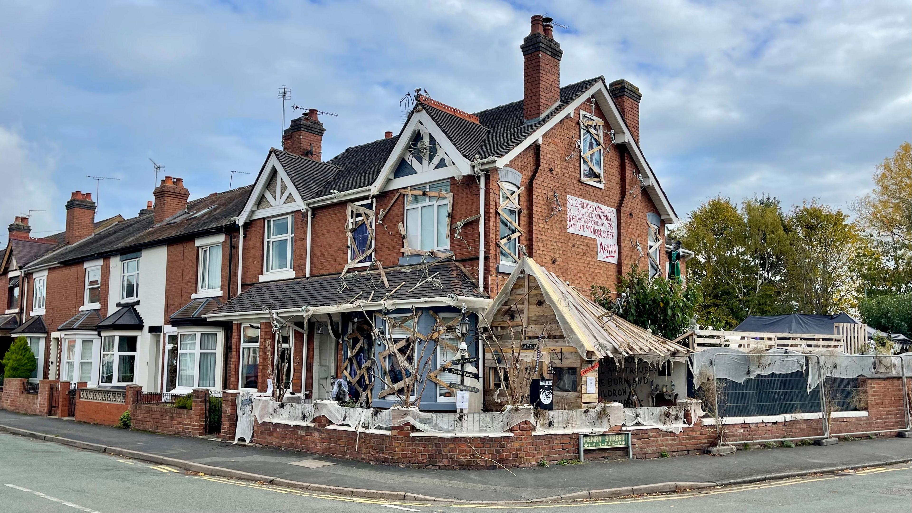 An end terrace house on the corner of a street covered in Halloween decorations. There are skeletons pinned to the wall and roof, planks of wood covering the windows, webbing across the fence and a huge wooden extension. Trees can be seen behind the house.