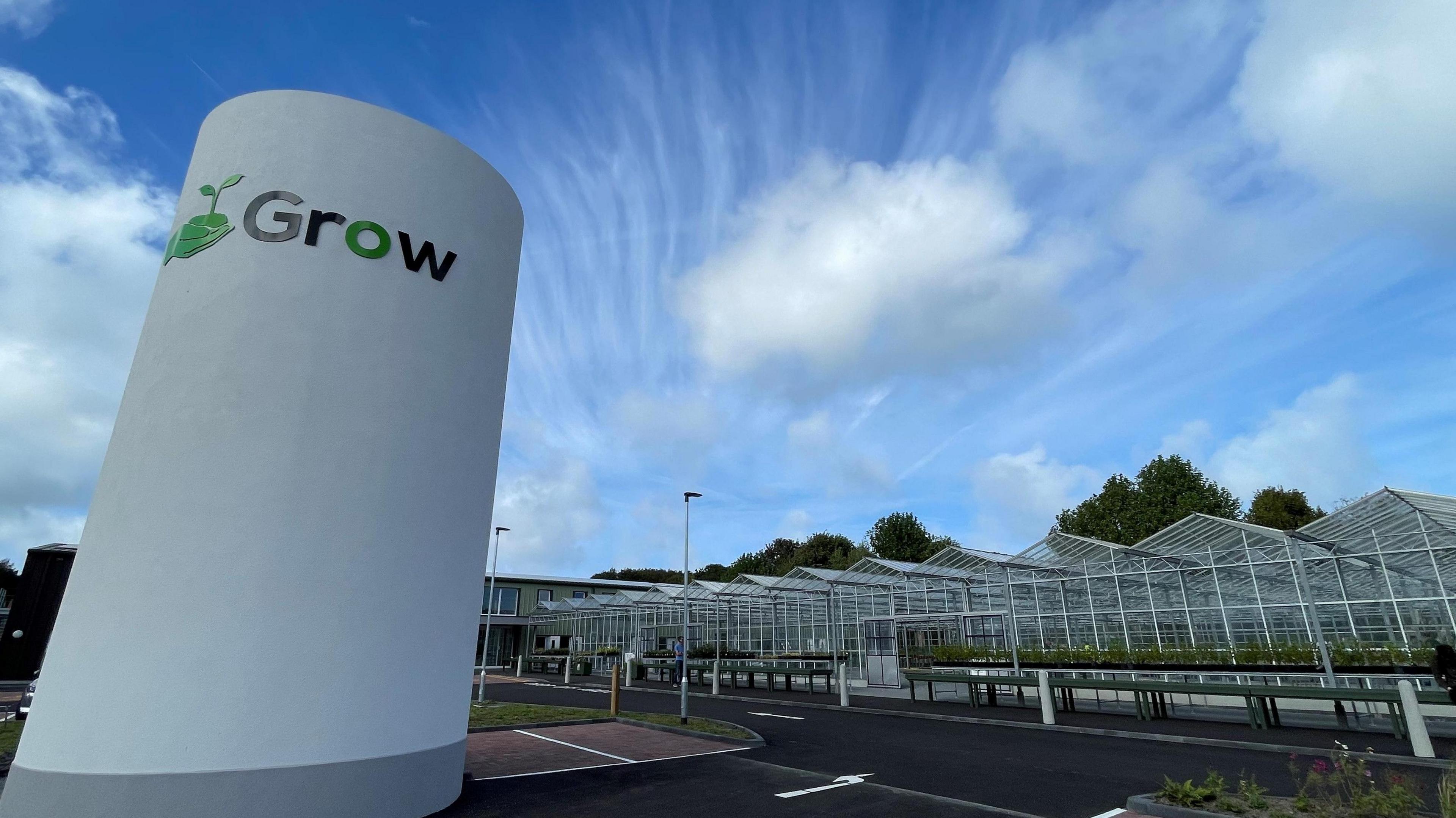 A view of the outside of the Grow premises. It shows a row of large glasshouses and a large white structure featuring the green Grow logo. 