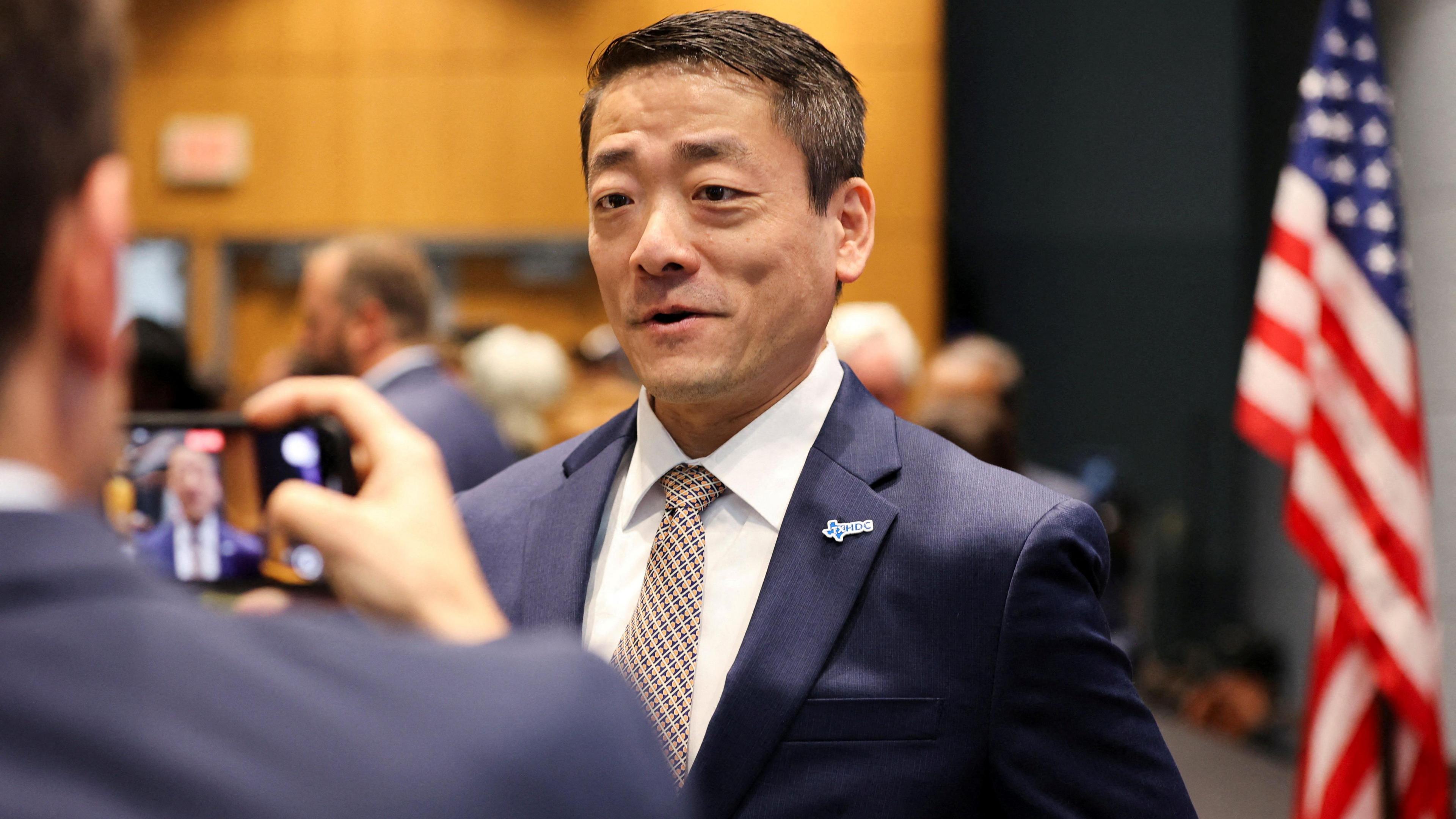 Texas lawmaker Gene Wu is seen in a suit in front of an American flag being recorded on another man's mobile phone.