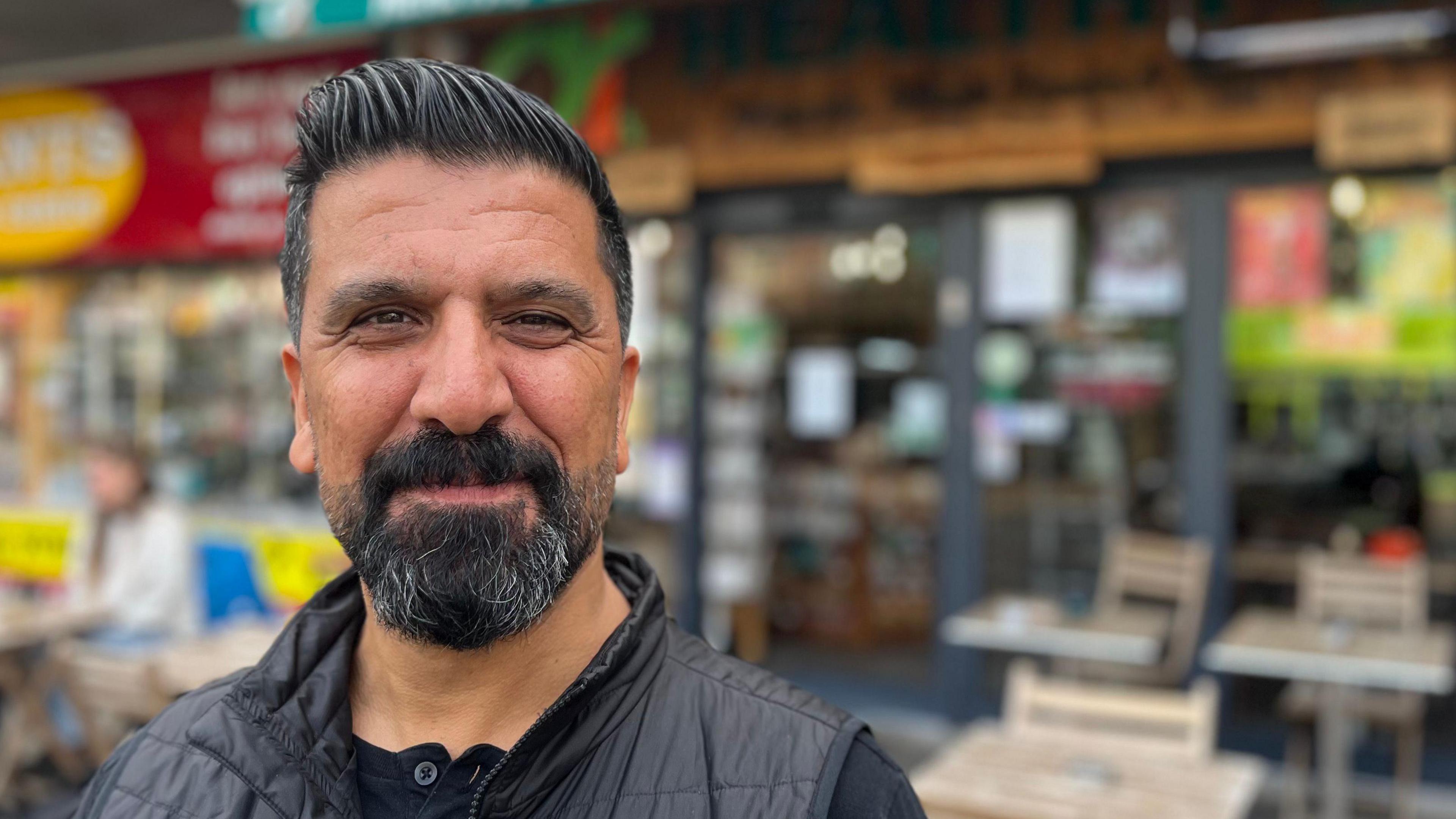 Mithat Ishakoglu wearing a black top and a black gilet in front of a blurred background of his health food shop and cafe.