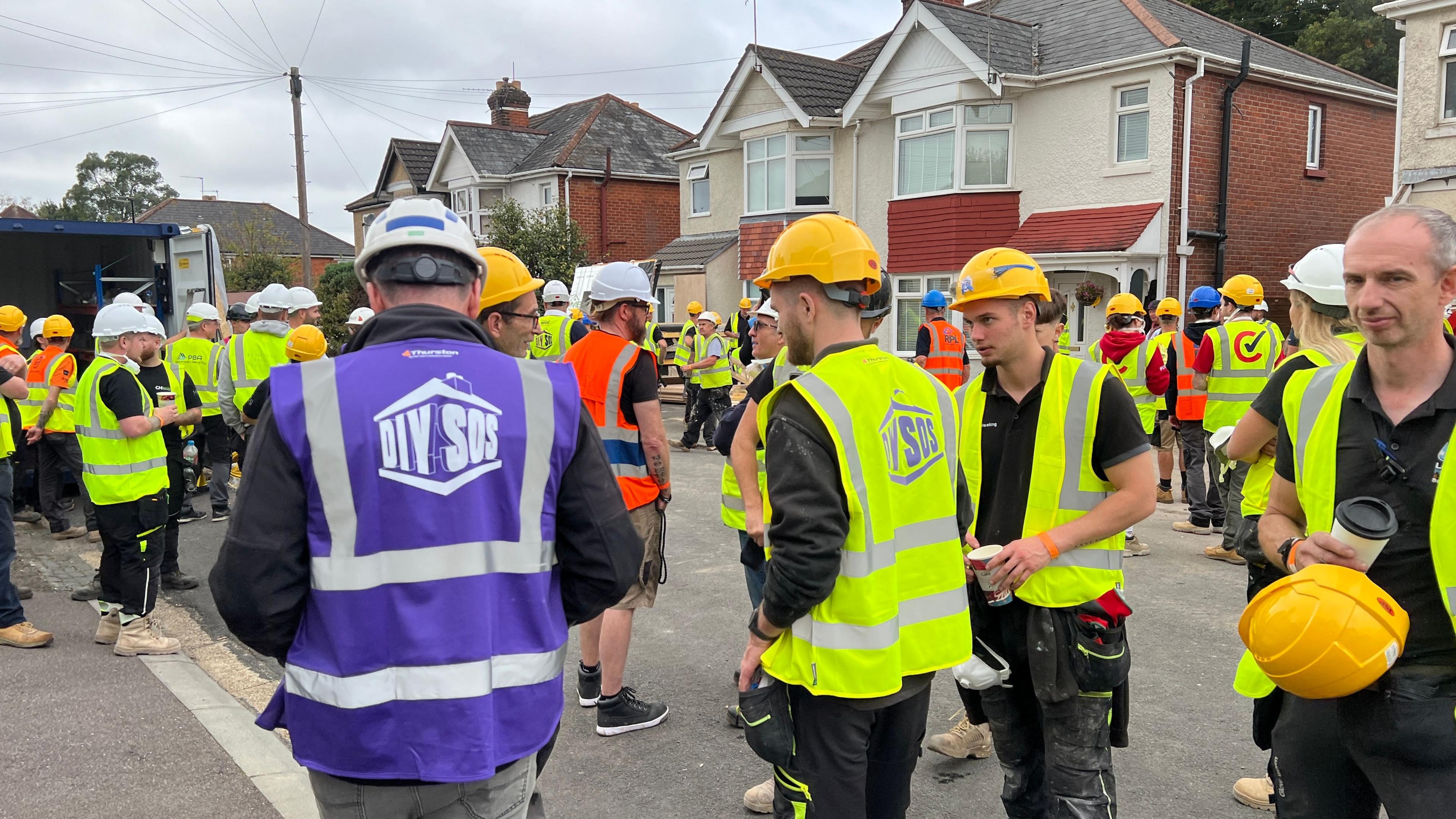 A group of men and women wearing florescent jackets and hard hats are standing on a street in front of a house where building work is being carried out. 