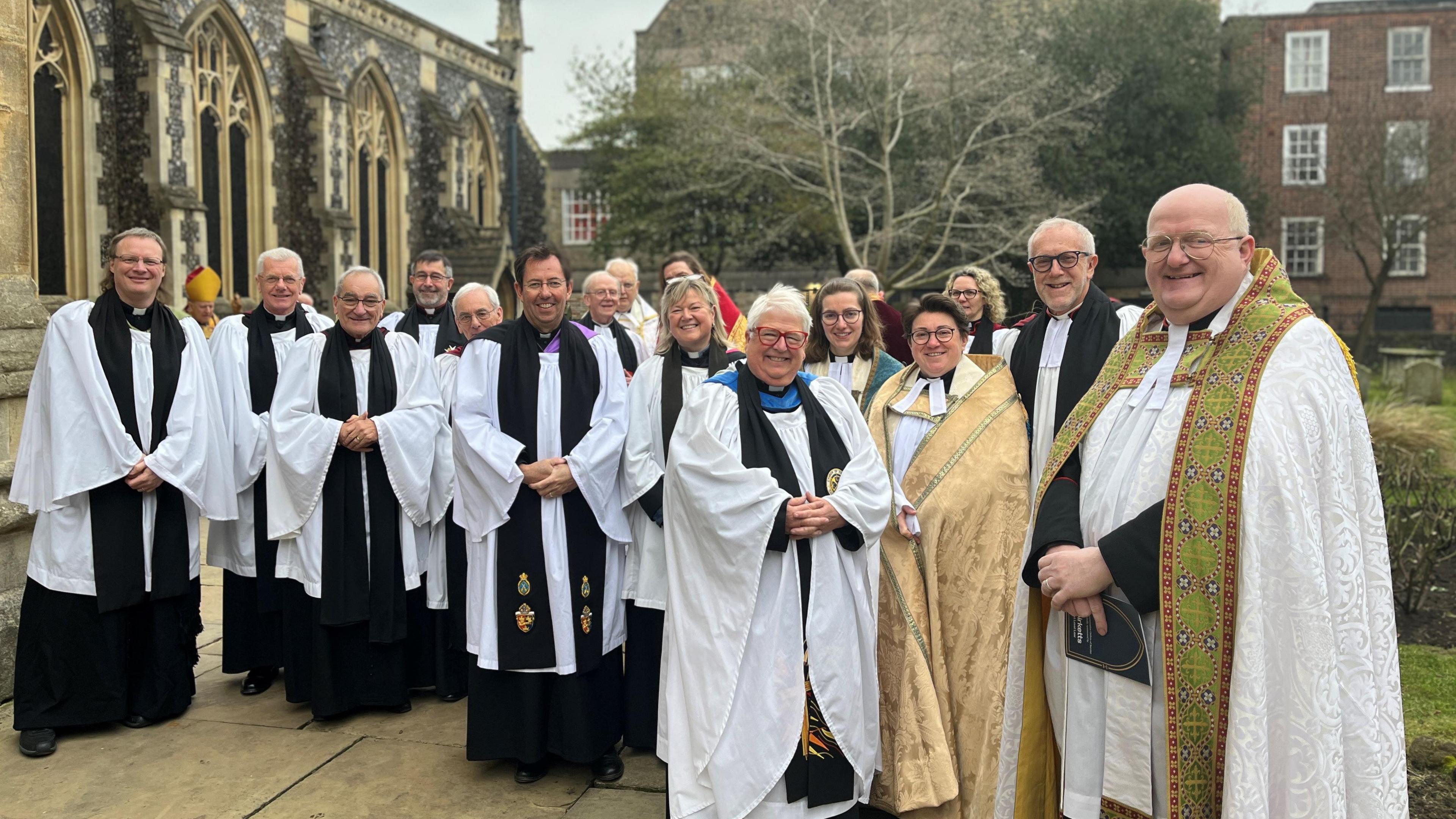 About 20 vicars in white robes stand outside a church.