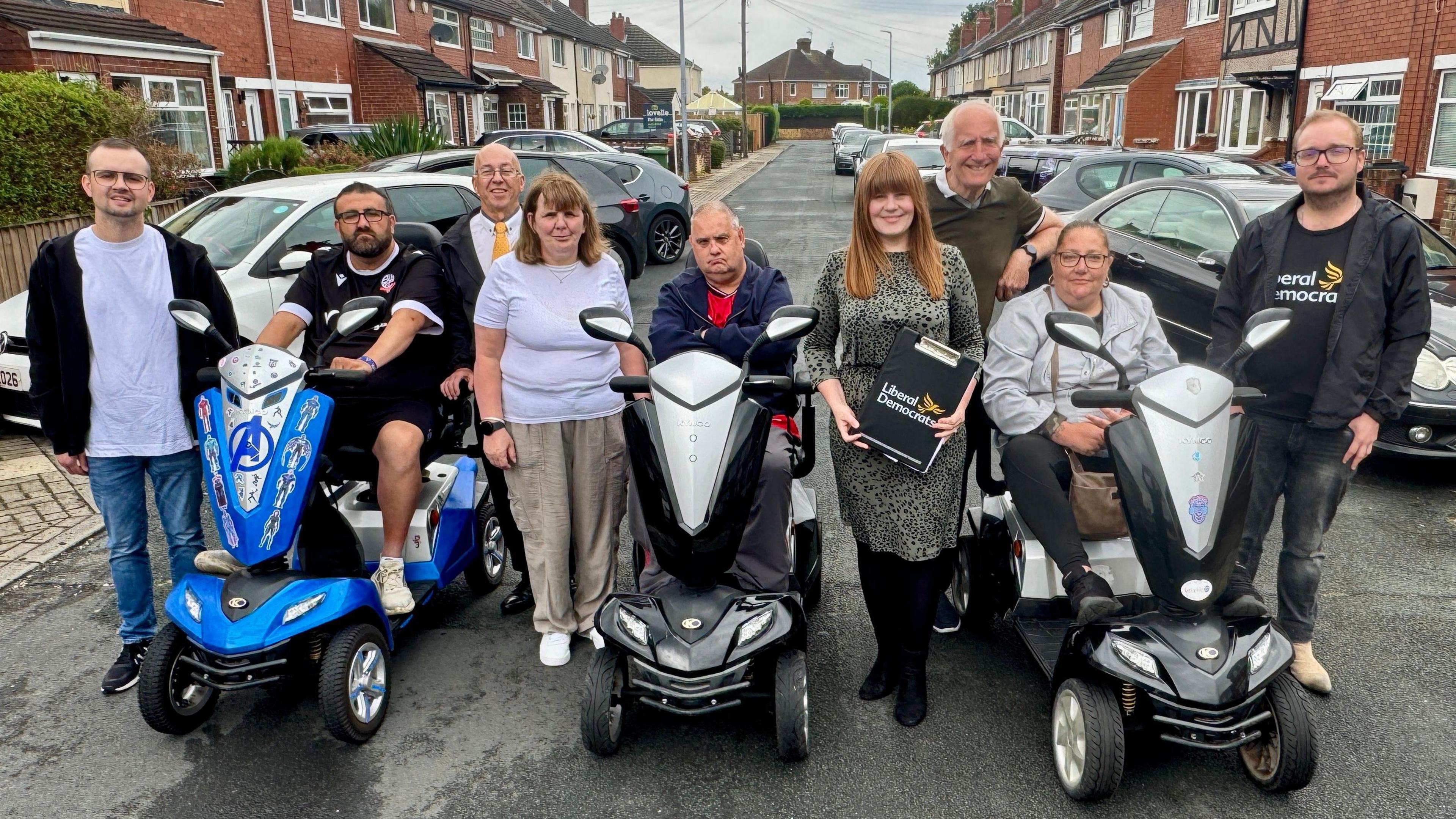Nine people in the middle of a road. Residential houses are on either side of the road and cars are parking in front of the houses on the road and paths. From left to right: A man wearing a black zip-up hoodie, a light grey t-shirt, blue jeans and black trainers. He has glasses. A man sat on a blue mobility scooter wearing a black t-shirt and black shorts. He has glasses and short brown hair. A man with glasses stood wearing a white t-shirt, a yellow tie and a black jacket. A woman with shoulder-length brown hair and a fringe. She is wearing a white t-shirt, beige trousers and white shoes. A man sat on a black mobility scooter wearing a red t-shirt and a navy jacket with his arms crossed. A woman in a knee-length green and black spotty dress holding a Liberal Democrats clipboard. She has long ginger hair with a fringe. A man with grey hair stood wearing a green t-shirt. A woman sat on a black mobility scooter wearing an off-white jacket, black leggings and black trainers. A man with glasses wearing a black Liberal Democrats t-shirt, black jeans, beige shoes and a black zip-up jacket.   