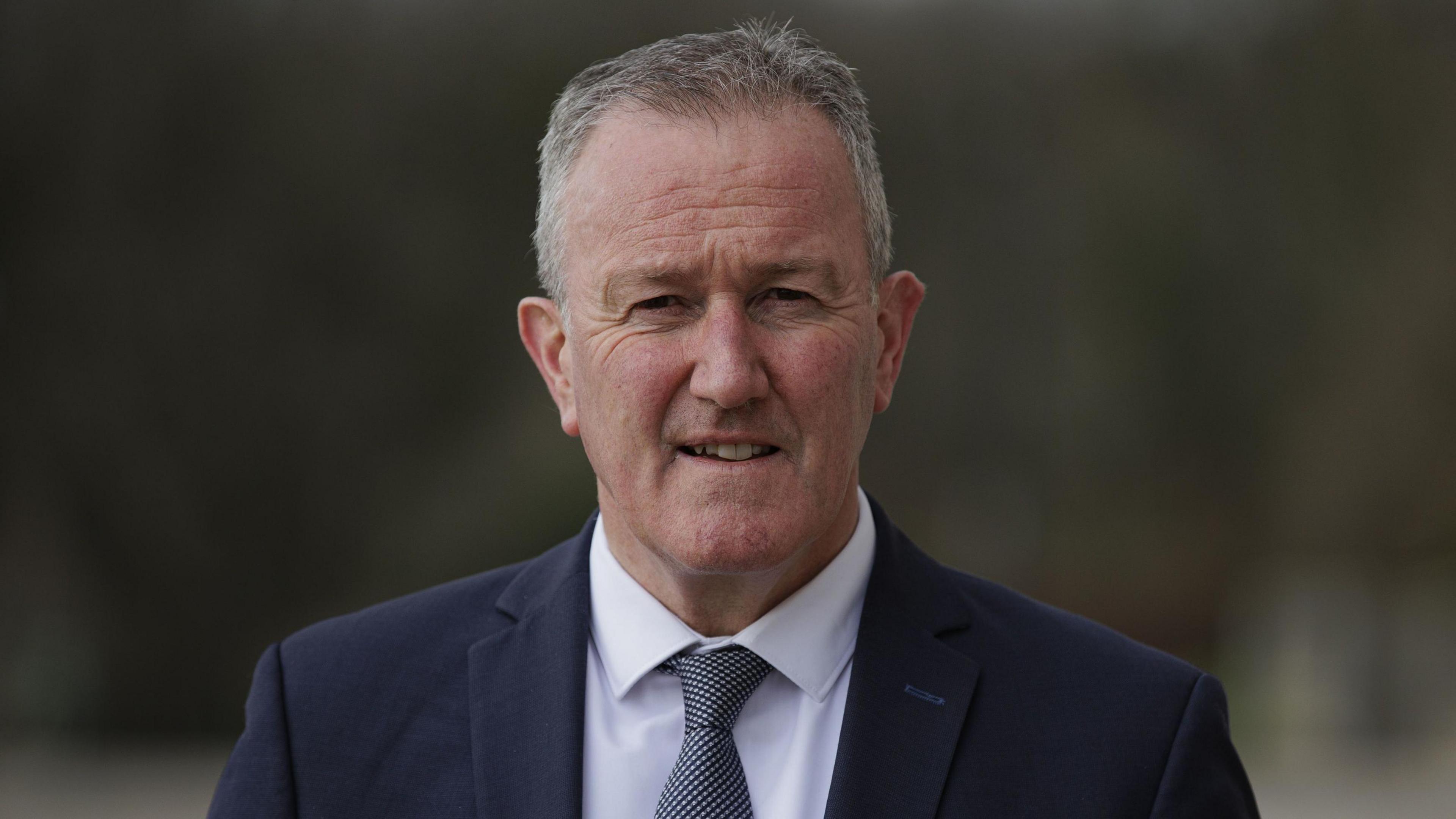 Conor murphy wearing a dark coloured suit, white shirt and tie. 