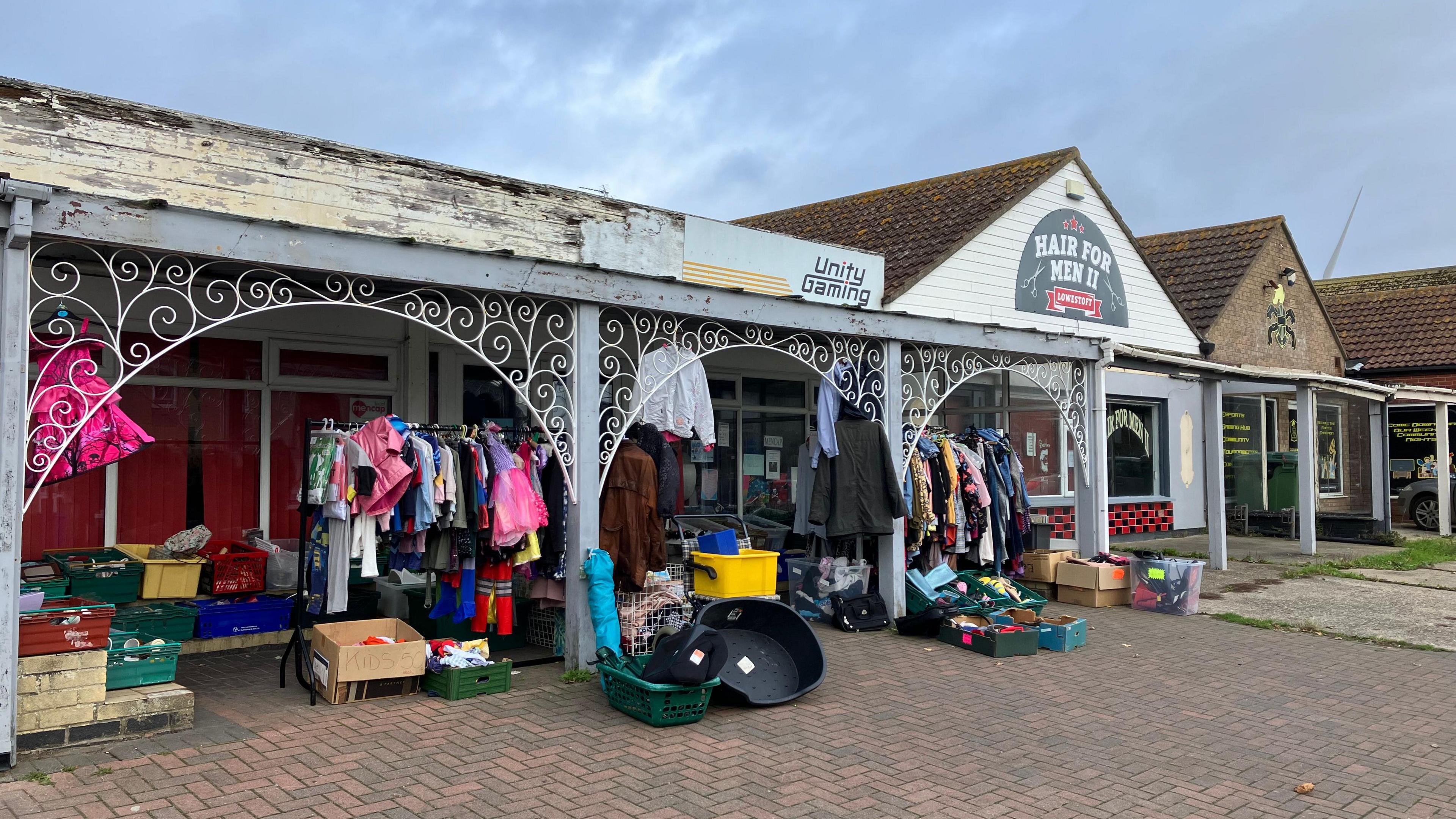 The exterior of a single-storey charity shop with clothes, dog baskets and colourful boxes with donated items  