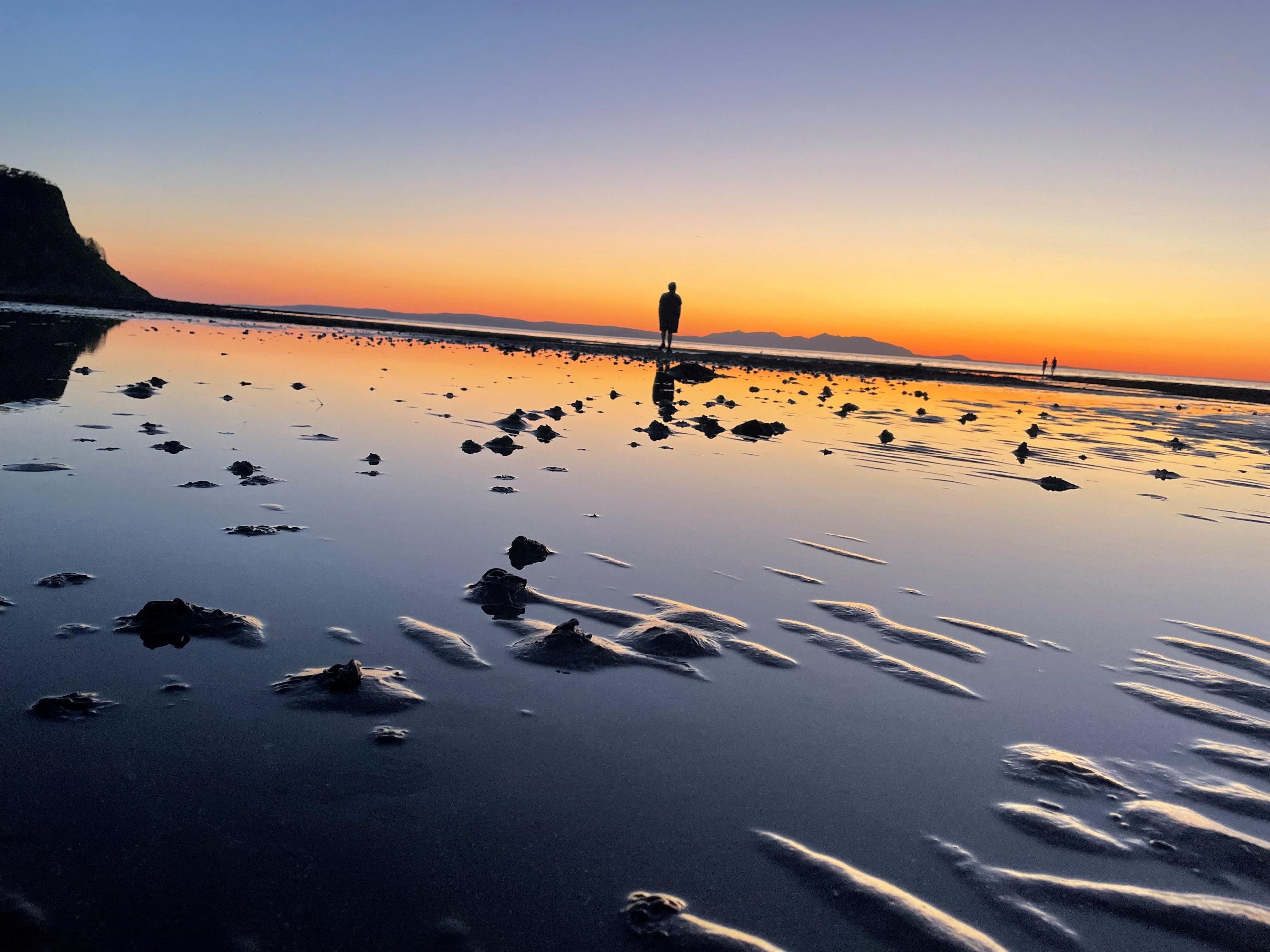 The sun sets at a beach, with a figure silhouetted in the distance