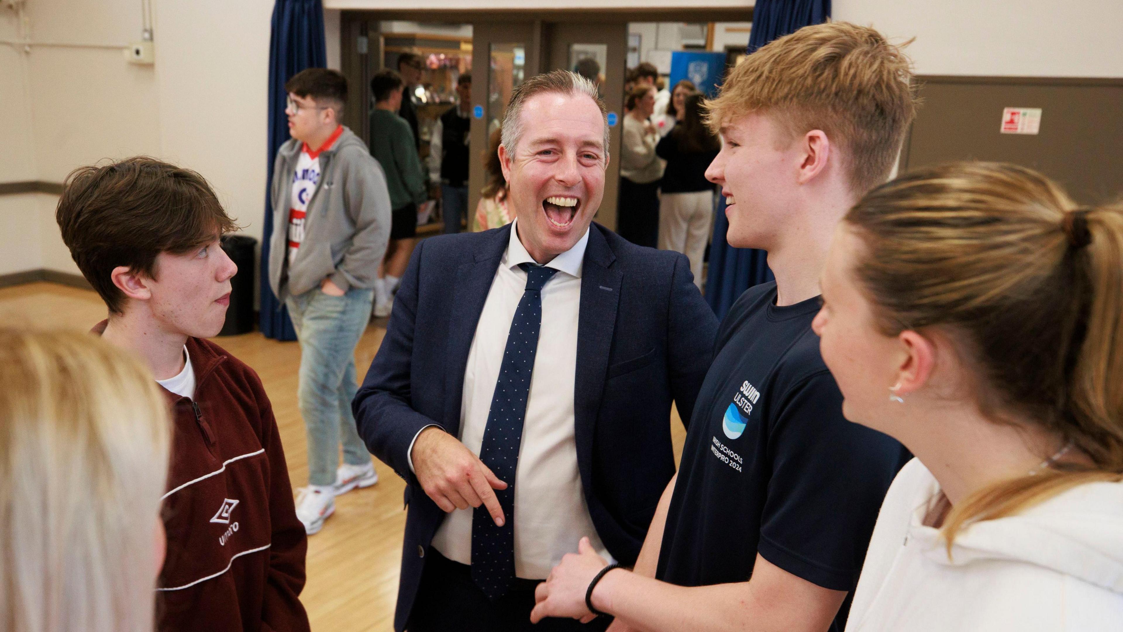 Education Minister Paul Givan smiling as he stands beside four young people in the hall of Belfast High School