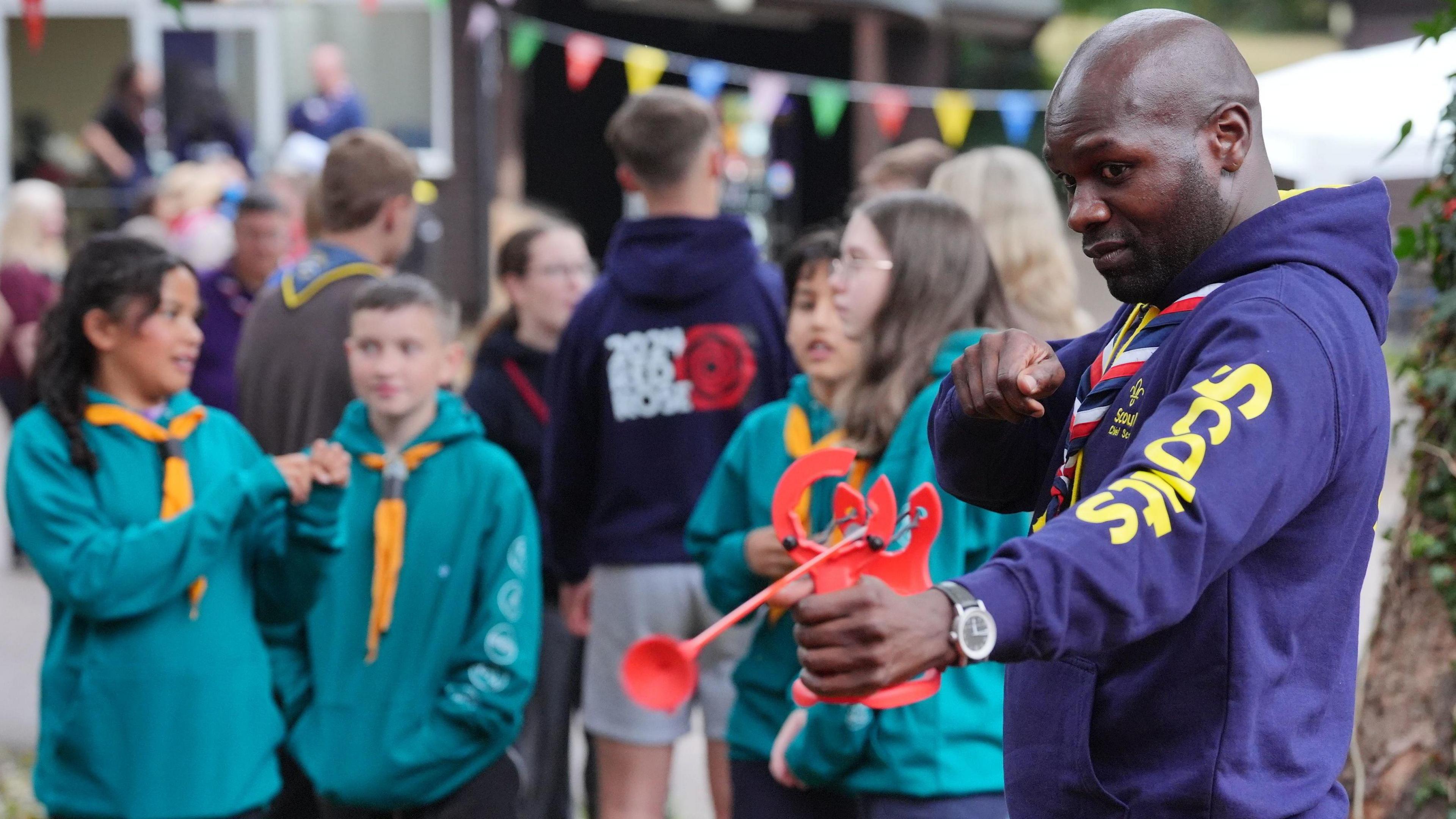 Dwayne Fields, wearing a Scouts hoodie and neckerchief, stands at the forefront of a lively outdoor event, aiming a red toy. Behind him, a group of children and young people in Scout uniforms chat and interact, with colourful bunting strung above them.