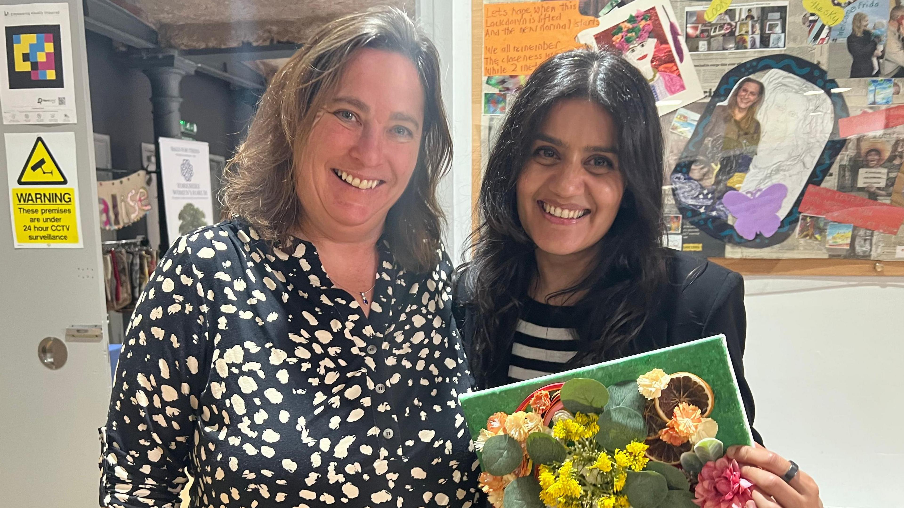 Two middle aged women stand smiling. One is white and has wavy, shoulder length brown hair. She wears a black and white spotty top. The other looks South Asian. She has long, straight, black hair and wears a black and white striped top and black jacket. She holds a piece of floral artwork.