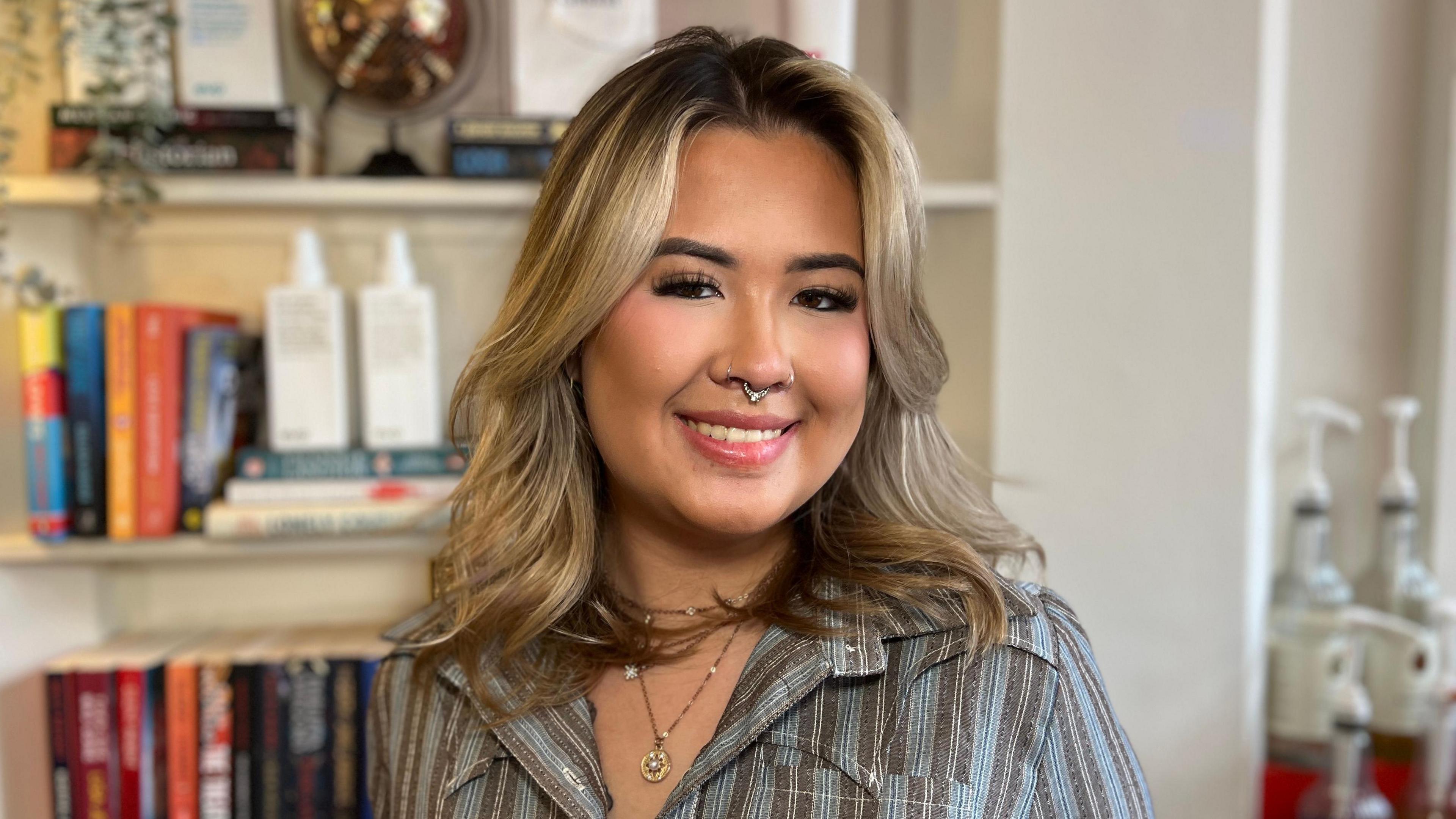 Natalie Bloor, a 21-year-old woman, with blonde wavy hair and dark roots. She is wearing a striped, collared shirt with layered necklaces. She has a septum piercing and is smiling.