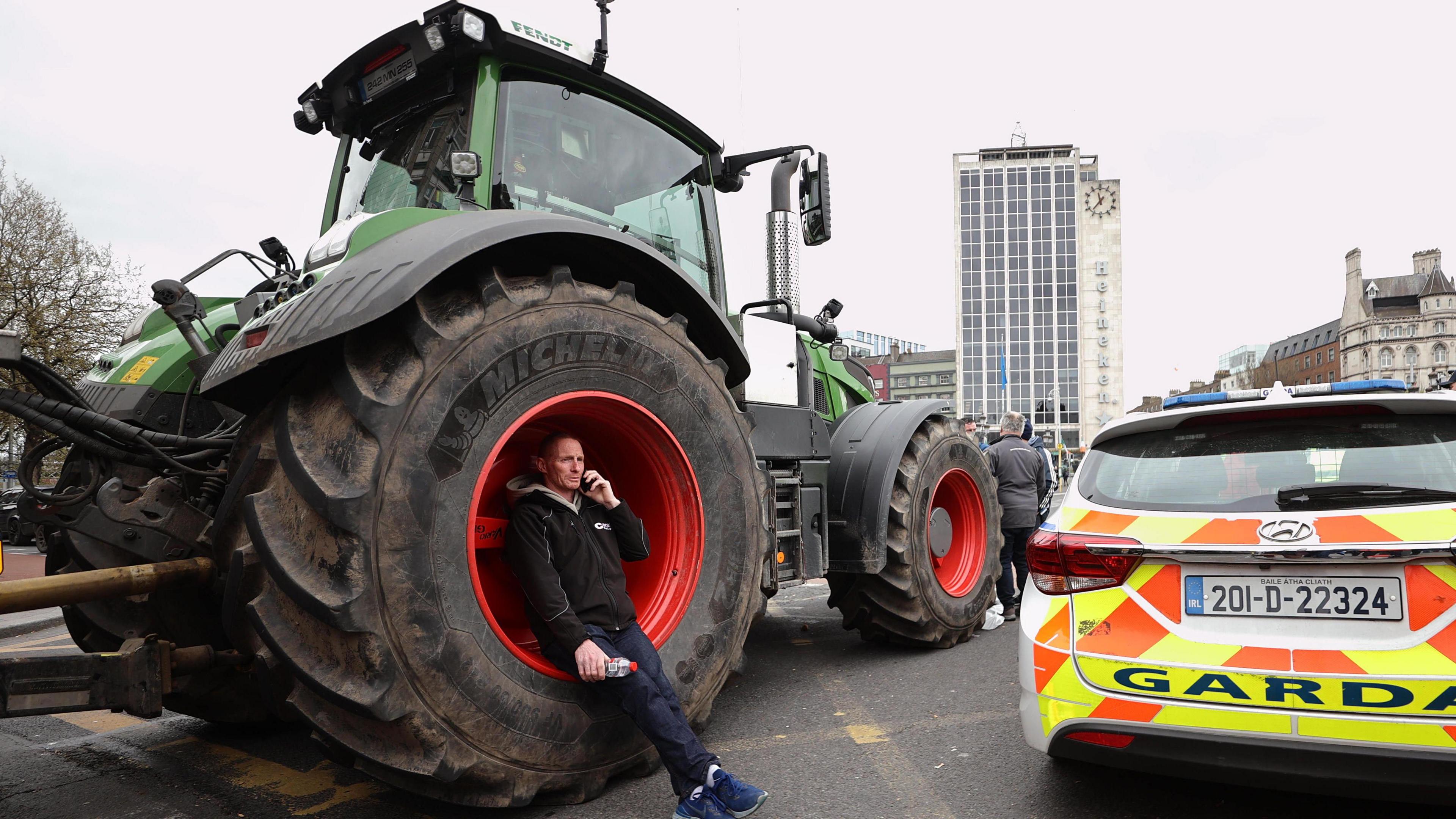 A man leans on a tractor tyre with a police car beside it that says GARDA