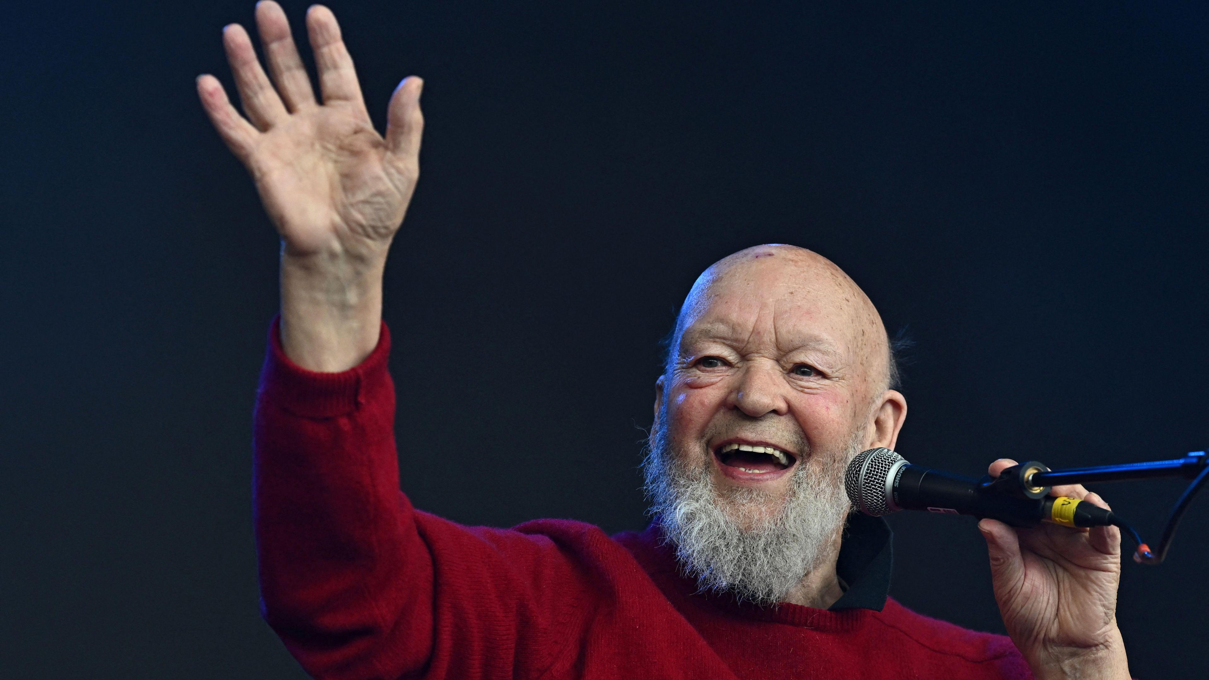 Michael Eavis is waving from a stage while holding a microphone. He is laughing. He has a grey beard and is wearing a red jumper.