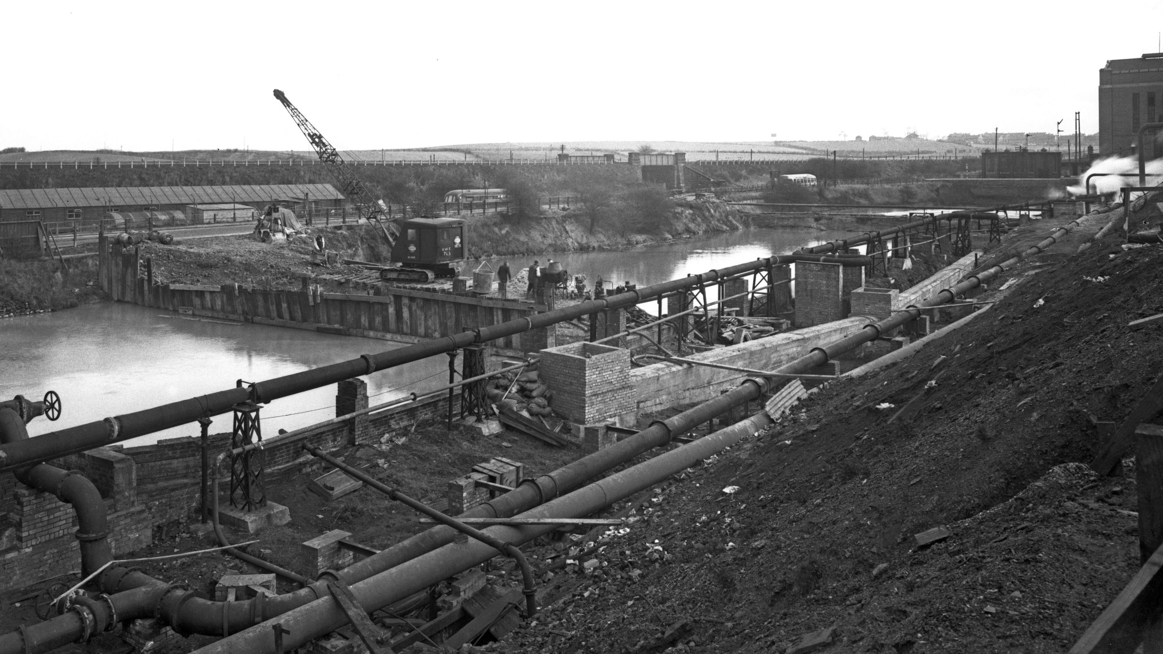 Construction work takes place on a mining site. Water and heavy machinery are visible on the photograph, which is monochrome.
