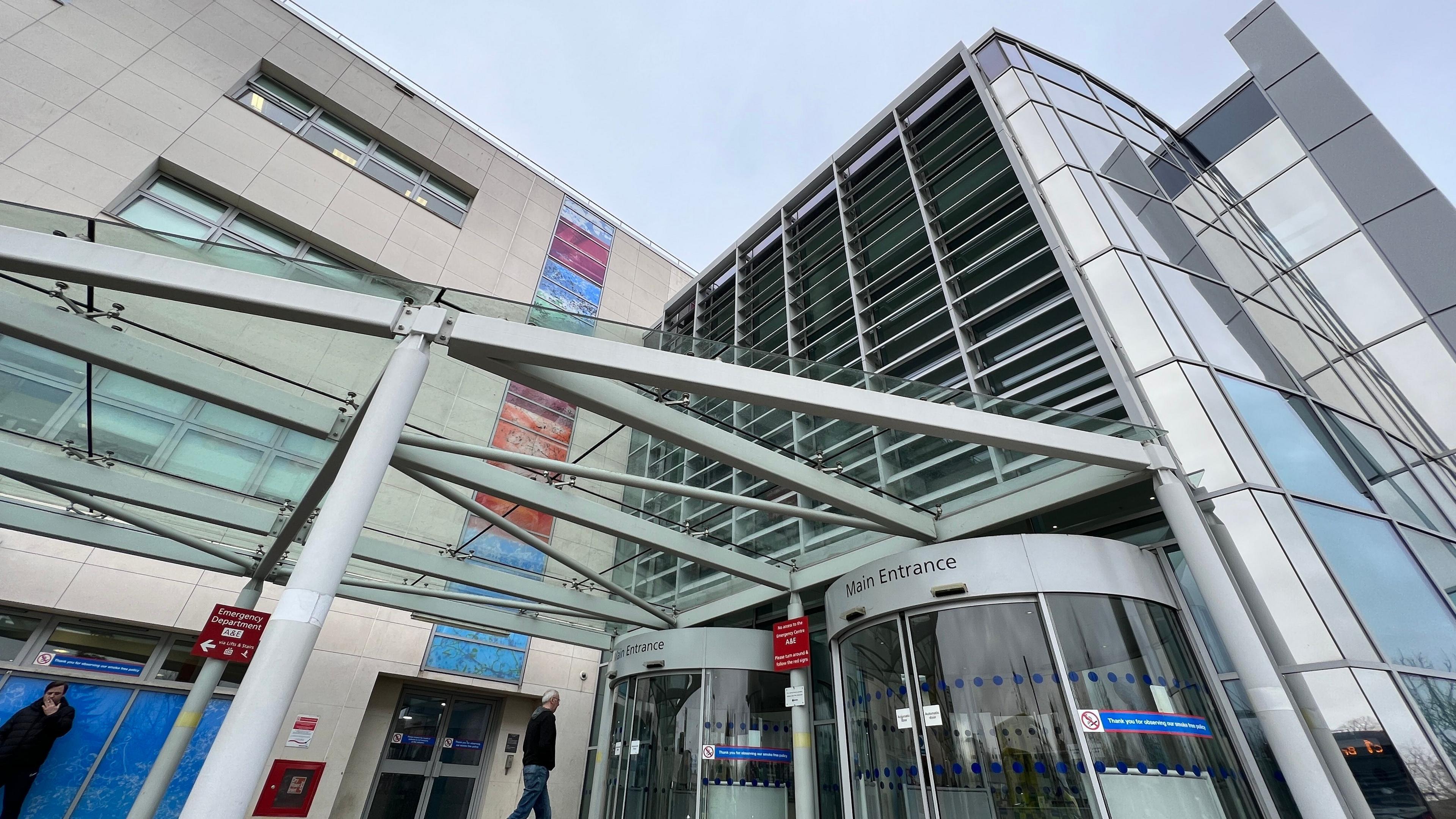 The concrete and glass exterior of a hospital shows a man approaching revolving doors with signs for the main entrance above them