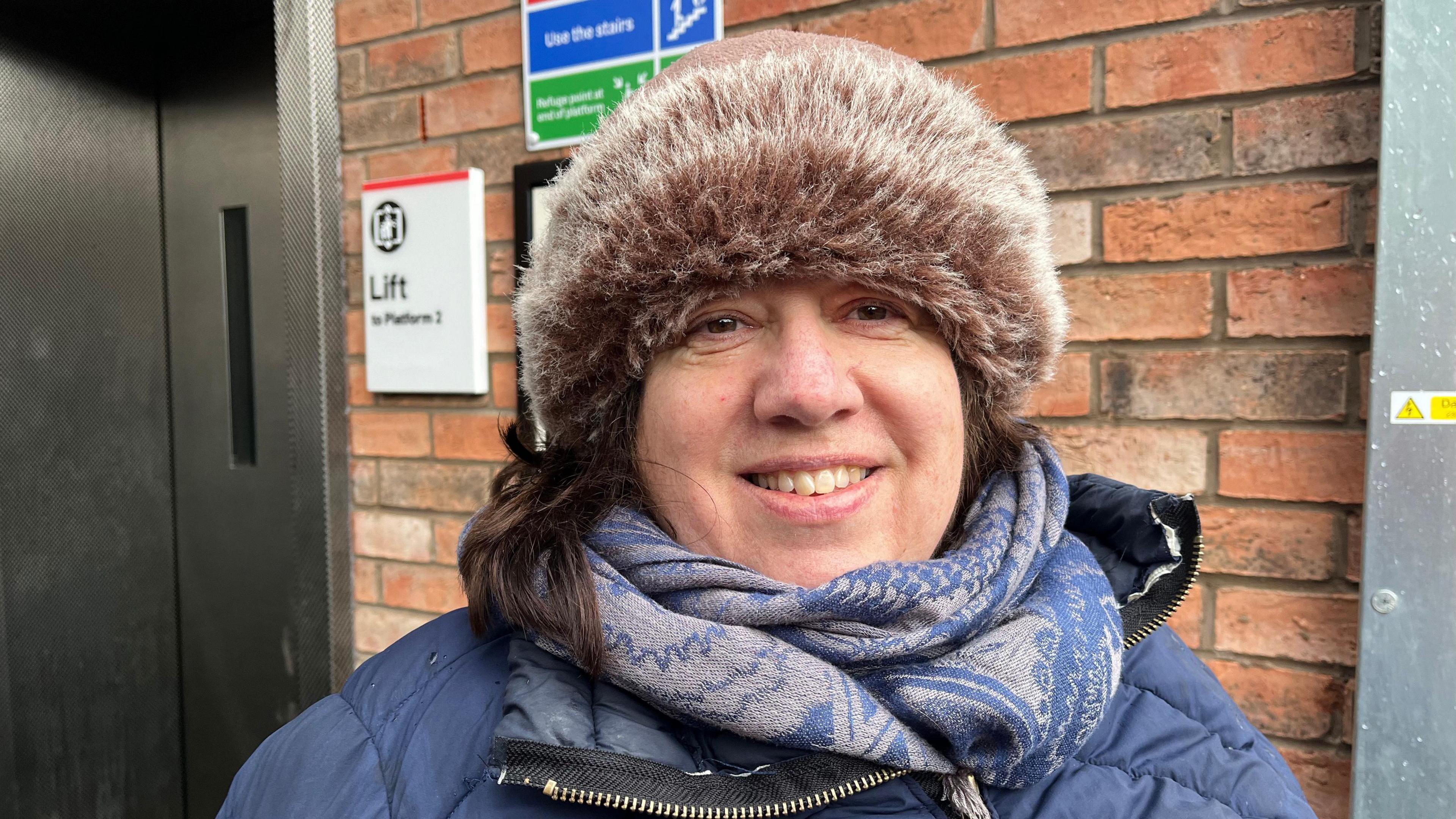 Amanda pictured looking into the camera and smiling. She's wearing a blue winter coat, a blue and grey patterned scarf, and a fluffy hat. Behind her is a red brick wall, with a sign to her right reading 'lift to platform 2.' Over her right shoulder, just out of focus are the silver metal lift doors. 