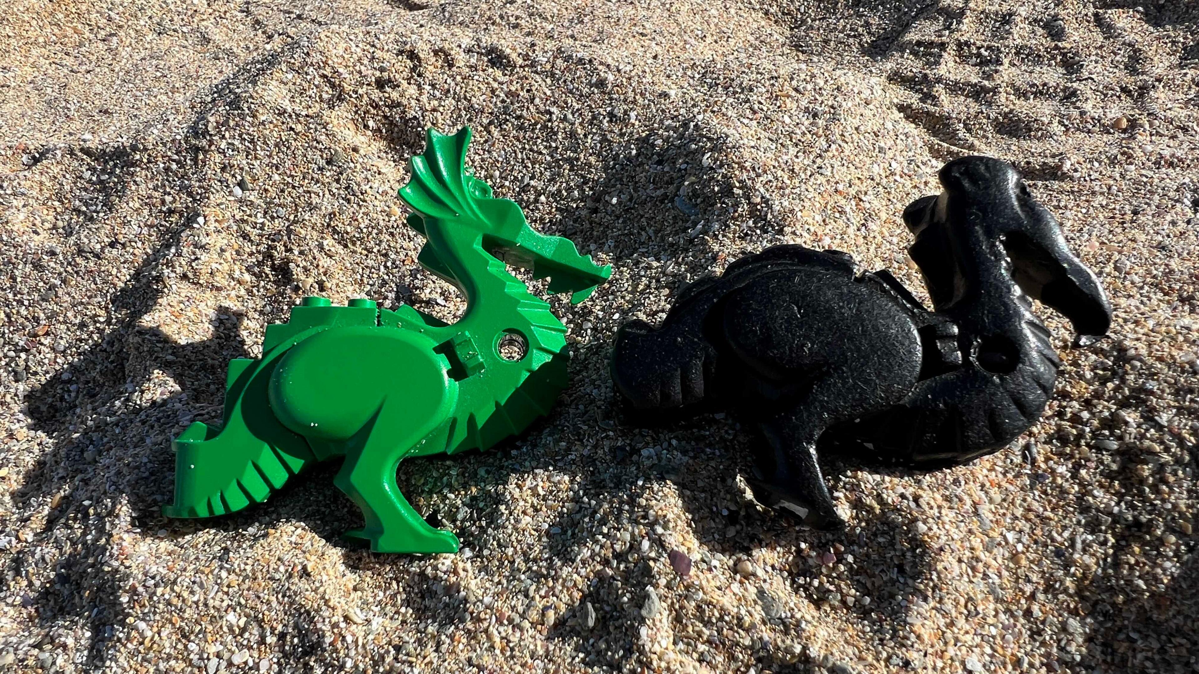 Two plastic dragon-shaped pieces placed on a sandy surface. One is green, featuring detailed wings, a tail, and a horned head. The other is black, also with intricate features like wings and a tail, but more worn.