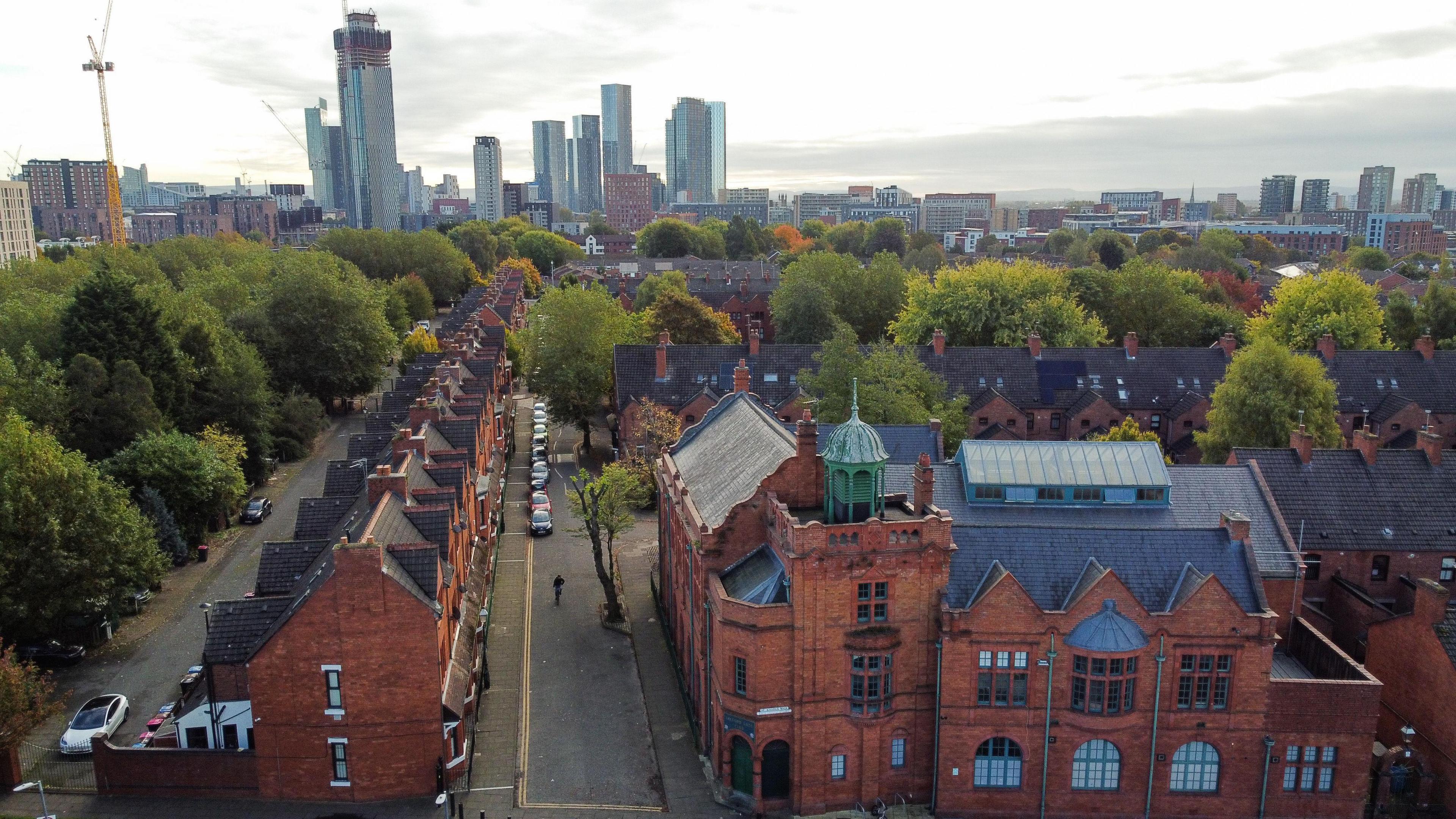 An aerial drone image of the Edwardian-built Salford Lads Club surrounded by old terraced housing and trees in the foreground, with skyscrapers and high-rises in the background