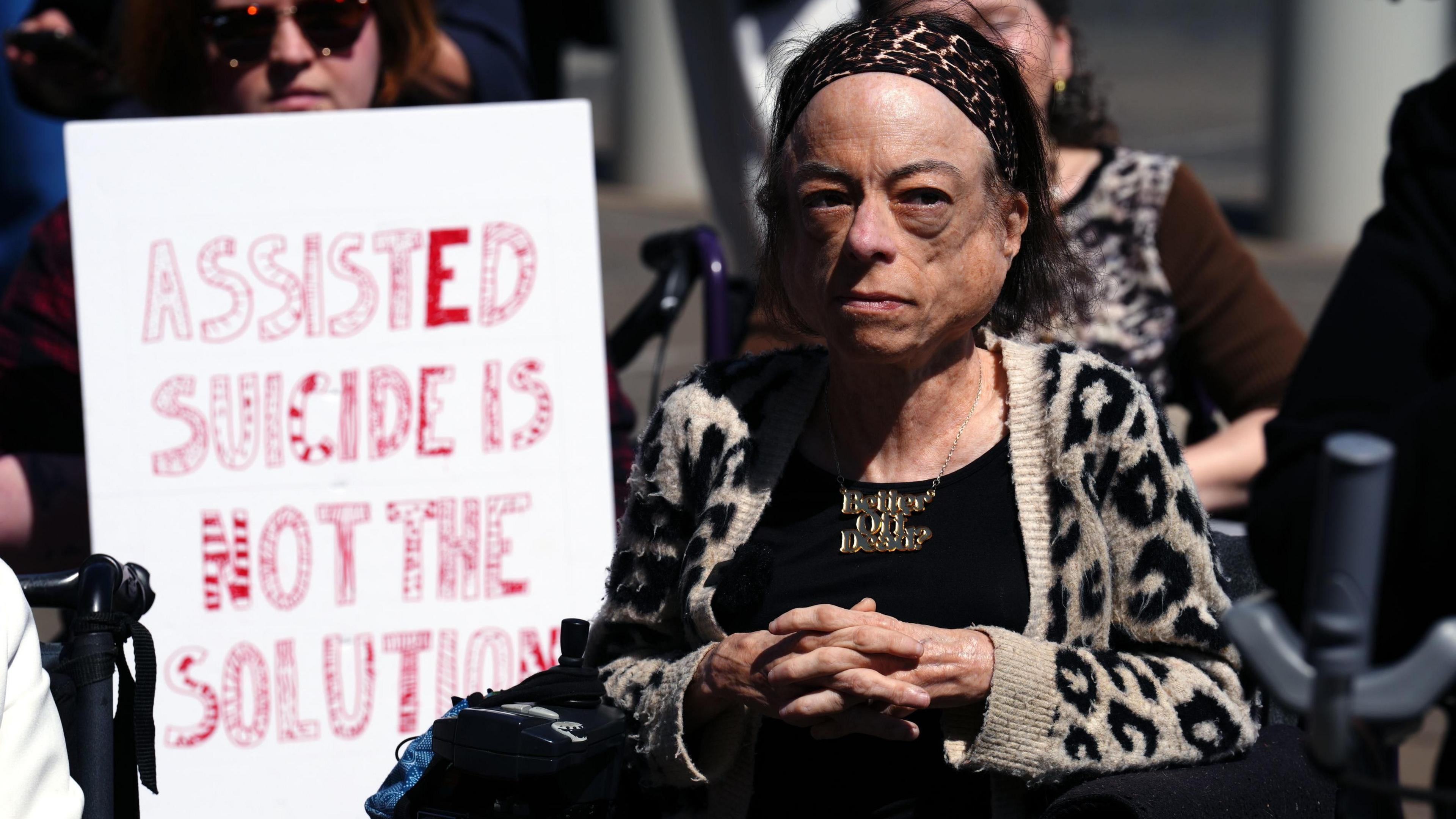 Actress Liz Carr demonstrating against the bill outside the Scottish Parliament. She is wearing an animal print cardigan and a headband, and a woman behind her is holding a placard saying: "Assisted suicide is not the solution"