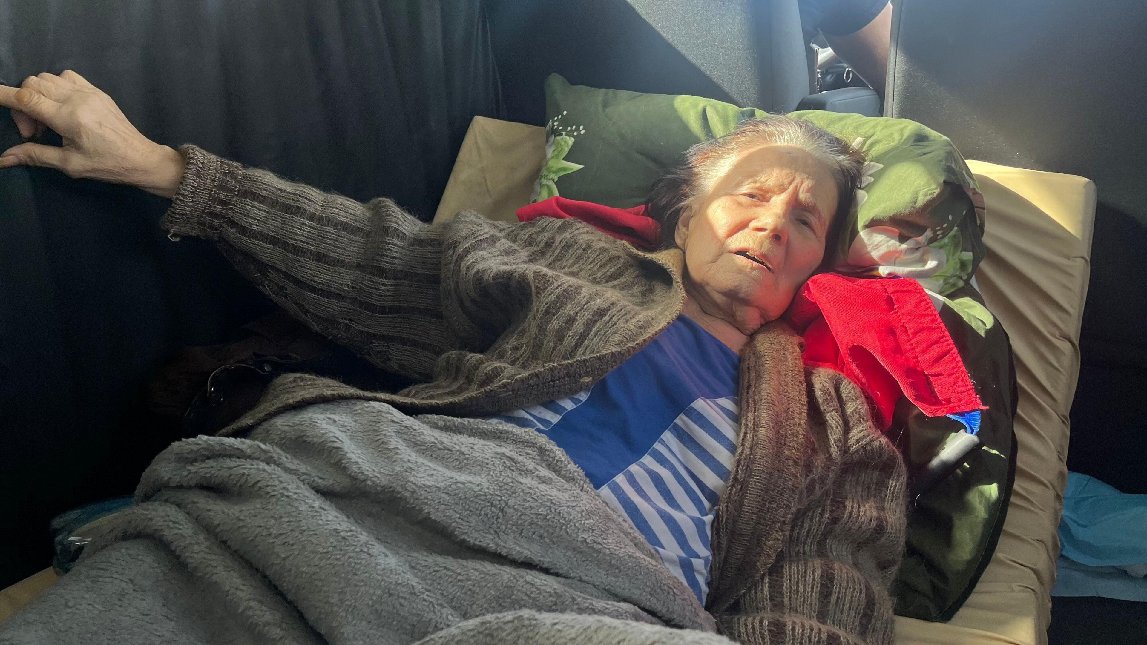 Nadiia lays on a mattress in a vehicle - her head on a pillow. She is wearing a brown woollen cardigan and a blue top. A grey rug is pulled up to her midriff.