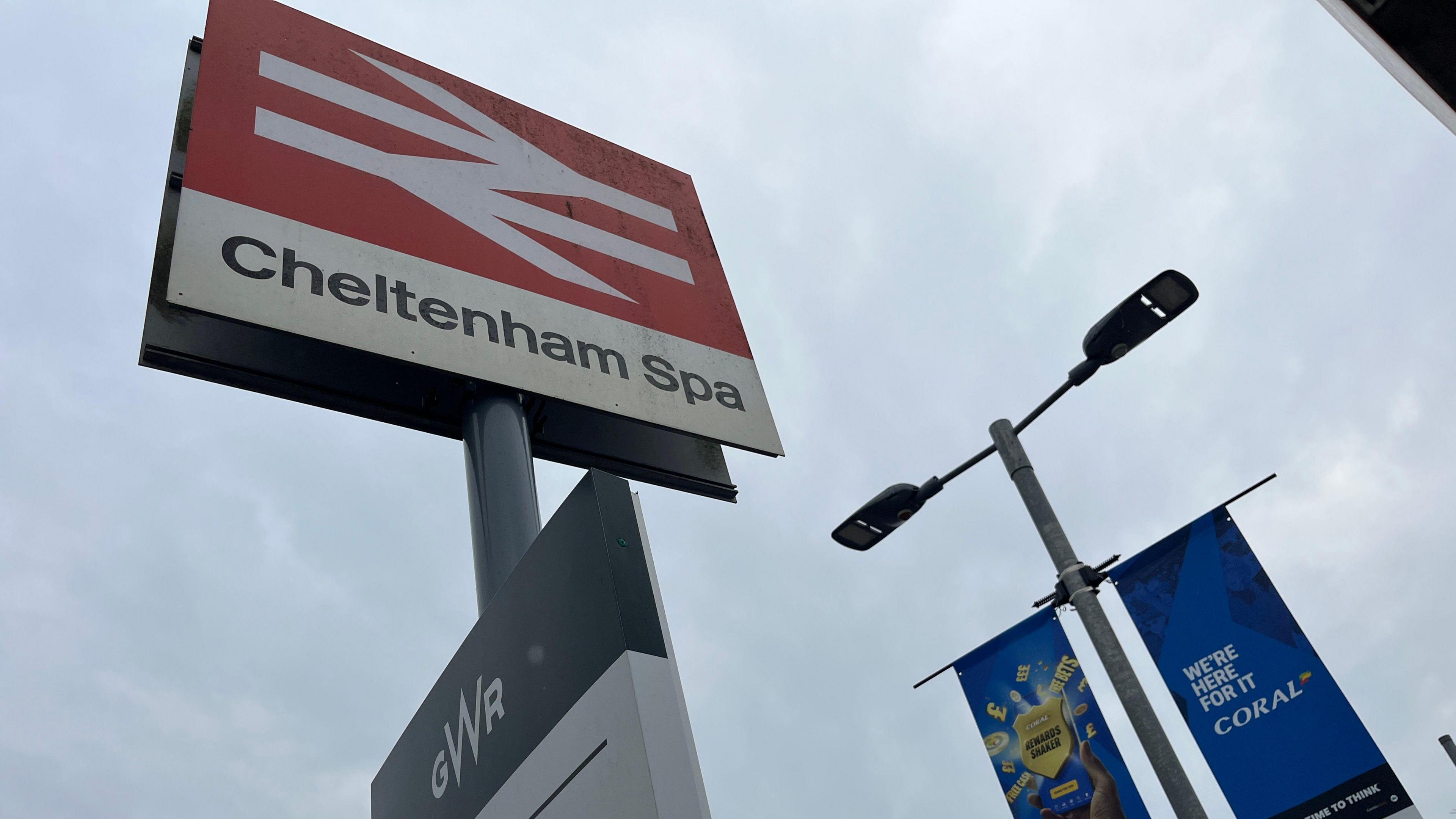 looking up at a sign that says "Cheltenham Spa" railway station on an overcast day.