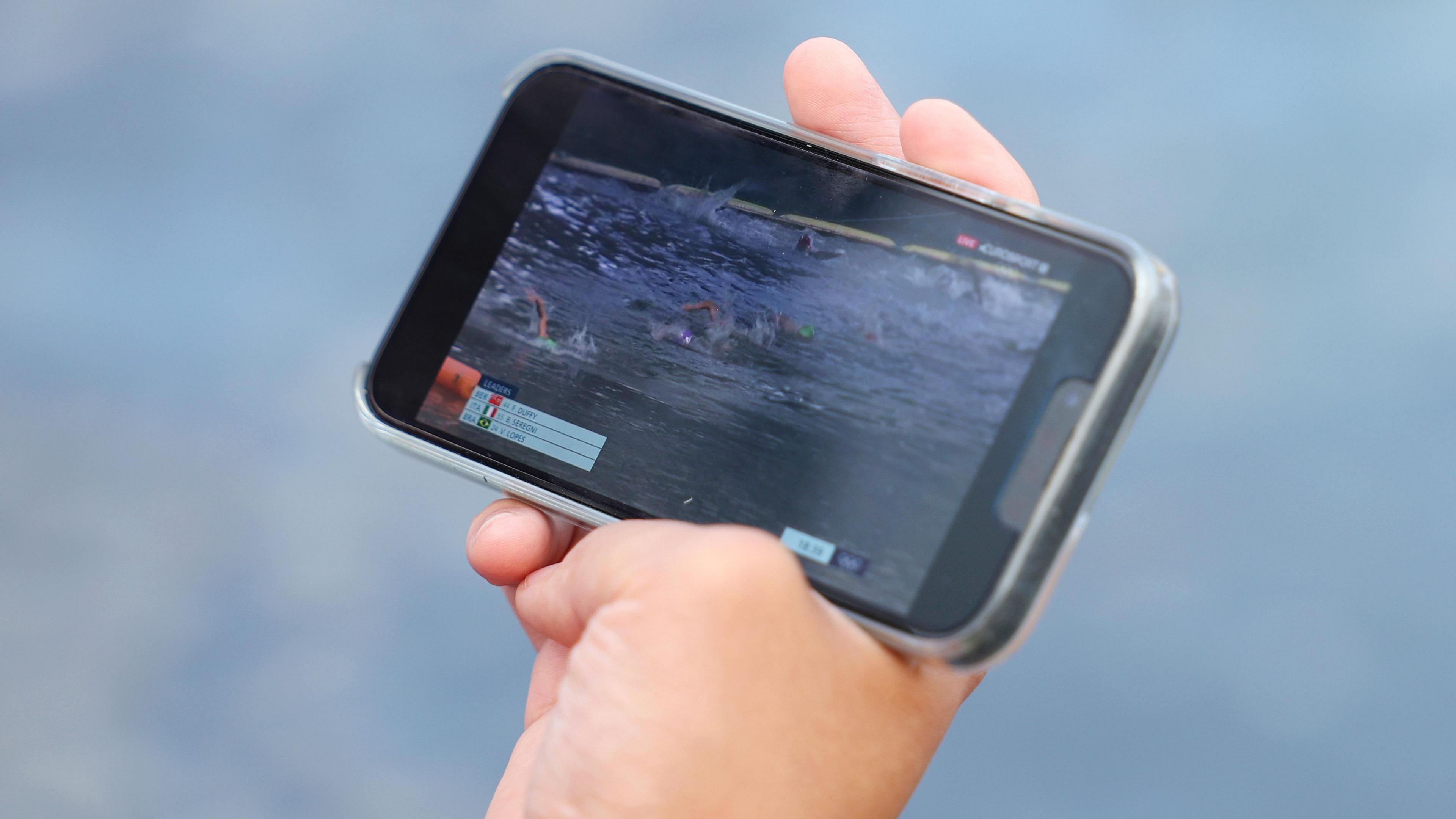 A person's right hand is pictured holding a mobile phone which is streaming video of the women's triathlon race at the Paris 2024 Olympics