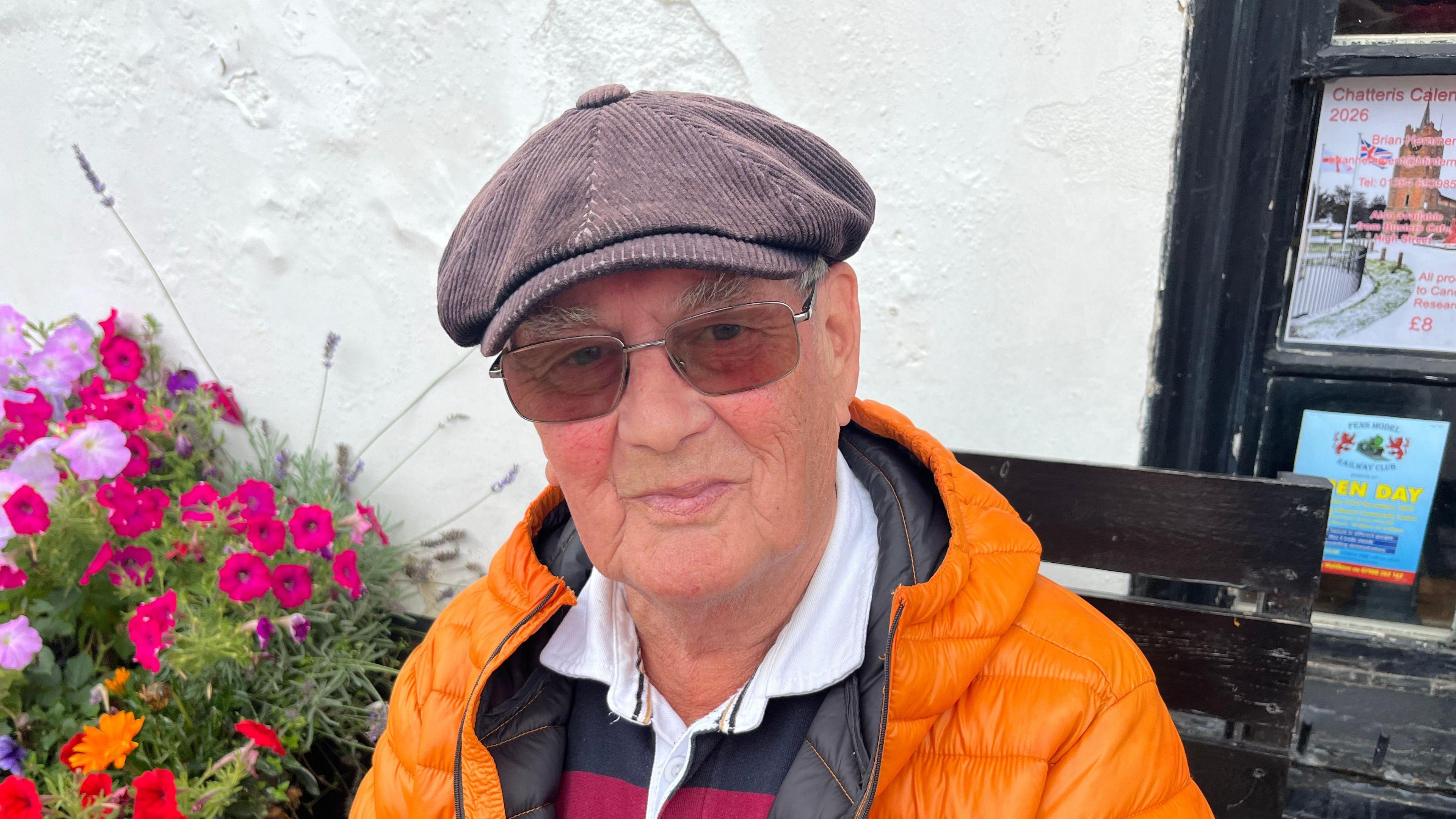 Trevor Colebrooke has a brown flat cap on and is wearing gold framed glasses with tinted lenses. He has a bright orange puffer jacket on and a rugby style maroon and black top on. He has quite bushy light brown eyebrows with a little bit of hair visible under his cap. He is sitting on a black wooden bench with a whitewashed building behind him. There are colourful flowers in a pot on the left.