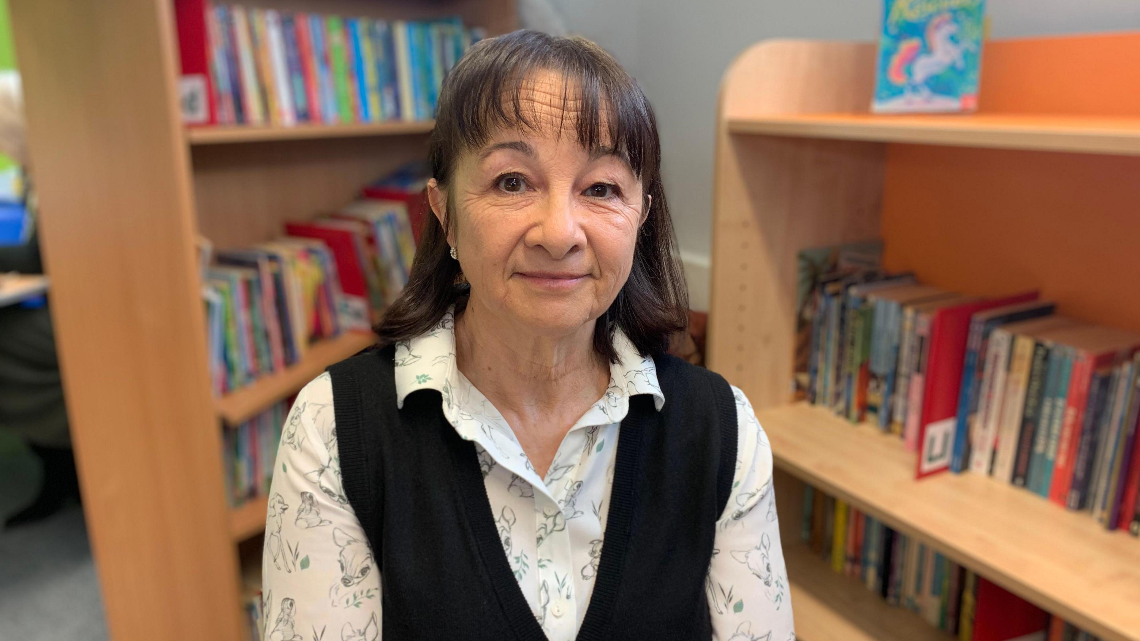 Speech and language therapist Sandy Chapell wearing a patterned top and glasses stood in front of a shelf in a school