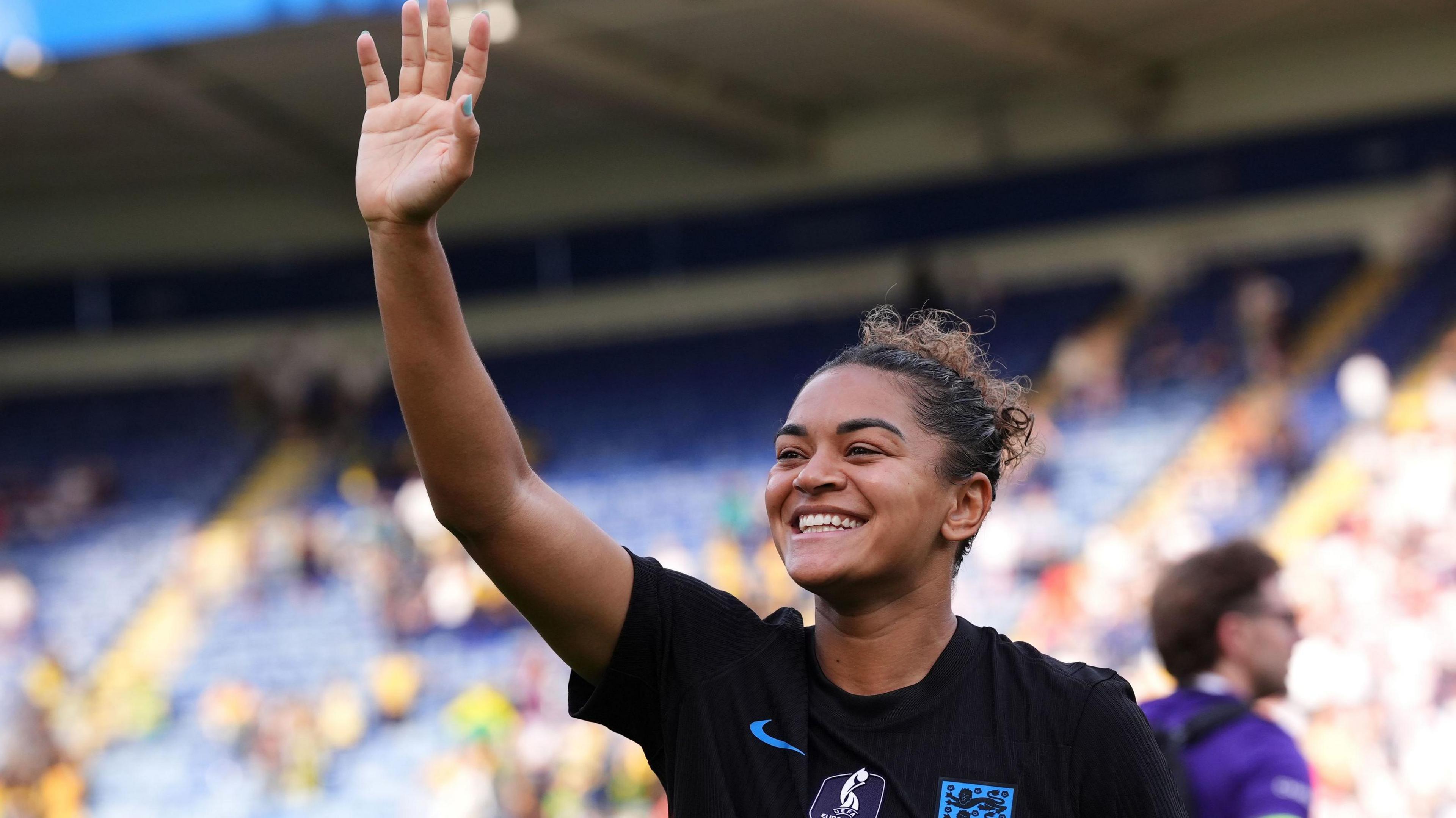 England defender Jess Carter smiles and waves at football fans. She is wearing a black England top with the club insignia on